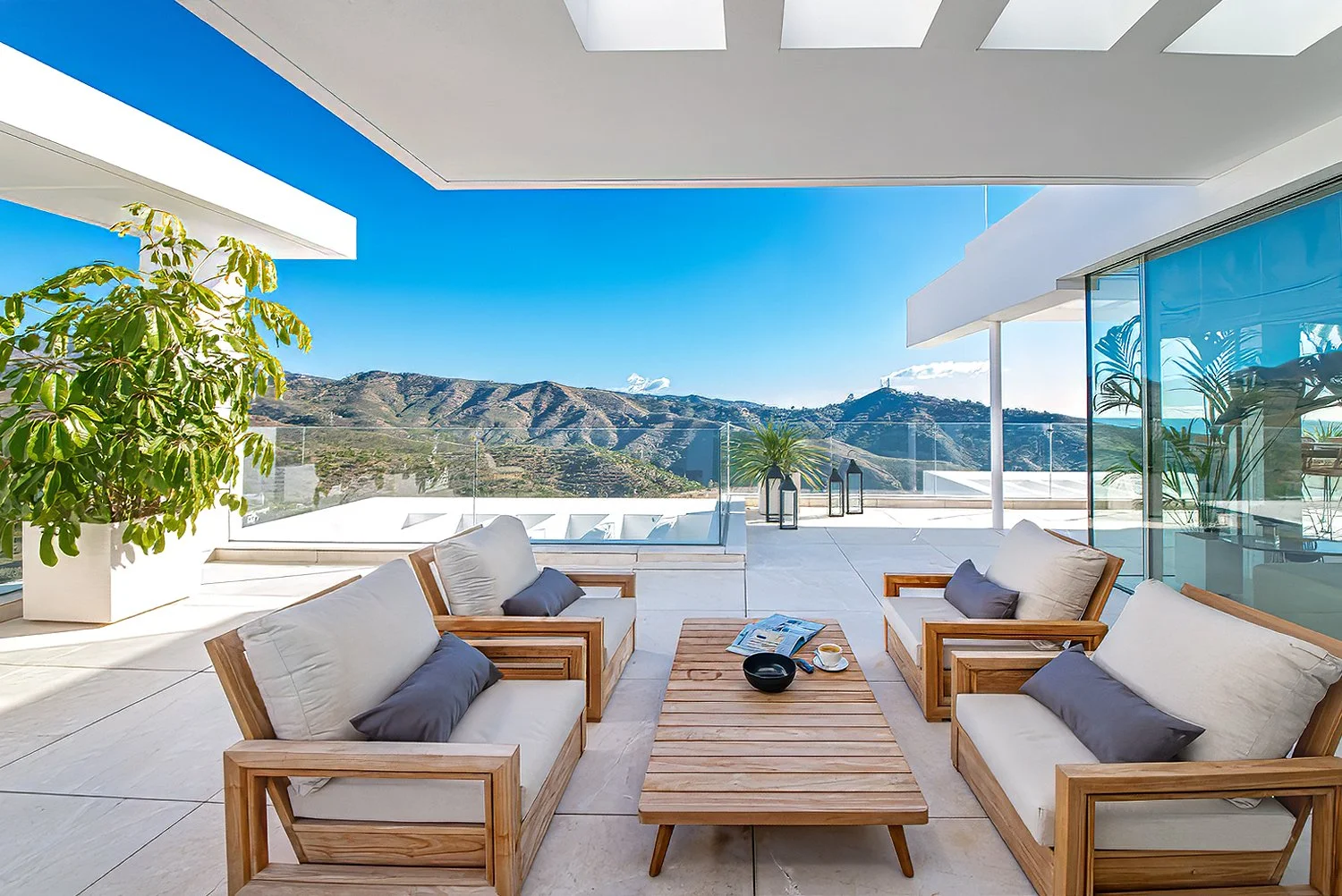Modern outdoor balcony with four wooden patio chairs with beige cushions and dark gray pillows, a wooden coffee table with magazines, a black bowl, and a cup, surrounded by potted plants and mountain views under a clear blue sky.