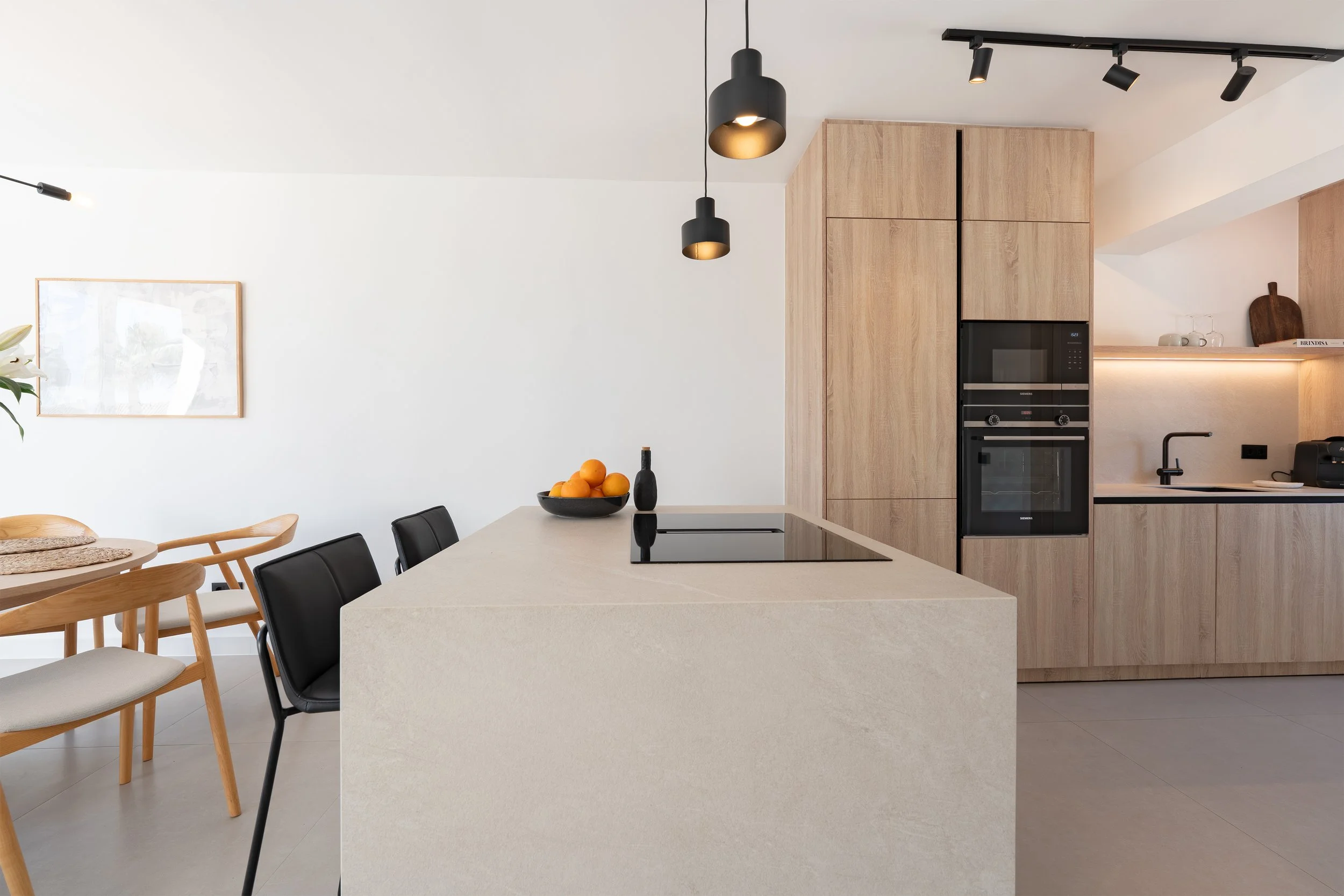 Modern kitchen with light wood cabinets, a black built-in oven, a black cooktop on a large beige island, and black pendant lights. There are black and wooden chairs around a dining table, a bowl of oranges, and a simple framed artwork on a white wall