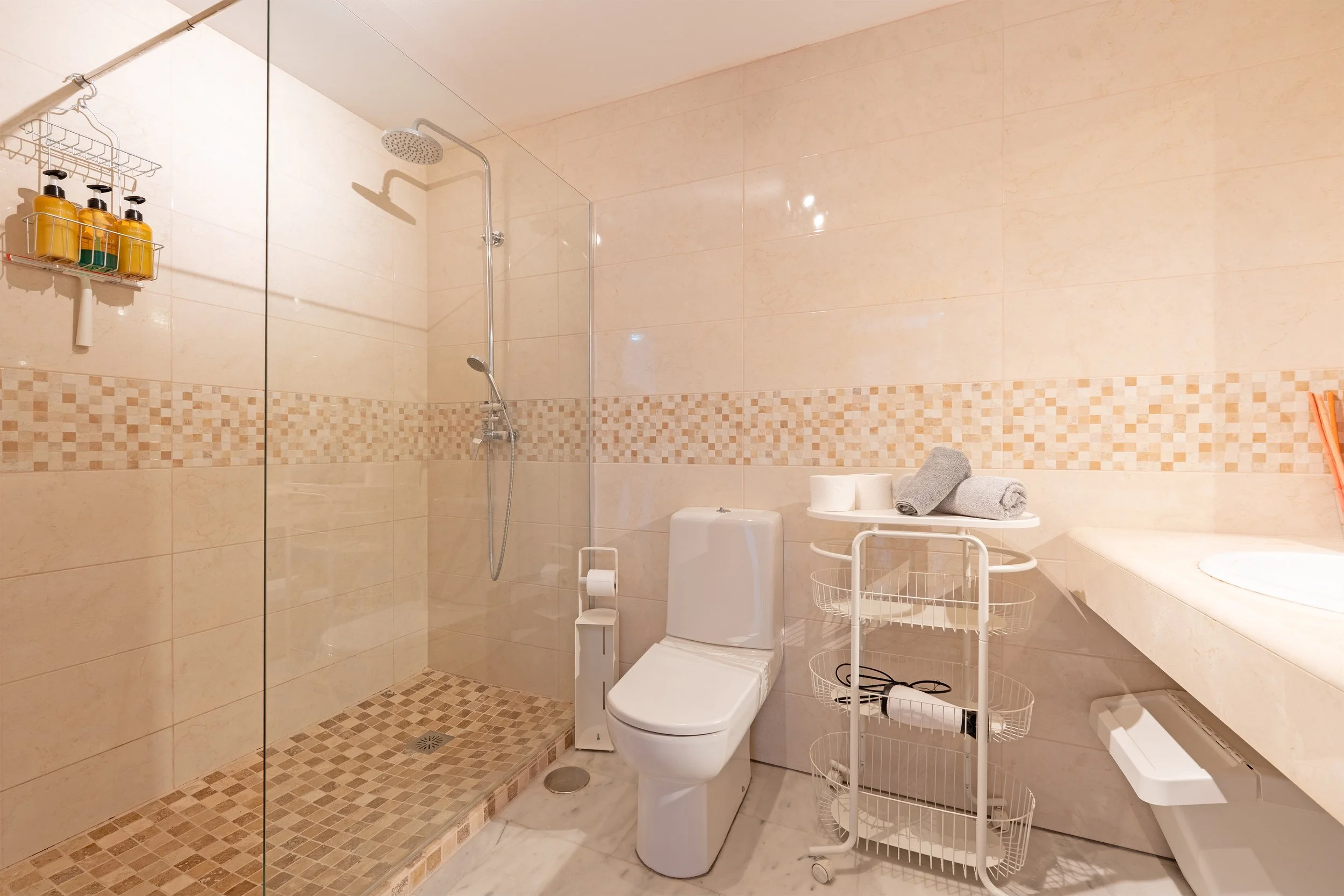 Bathroom with a walk-in shower with a glass partition, a white toilet, a small rolling cart with towels and toiletries, a marble countertop, and wall tiles in beige tones.