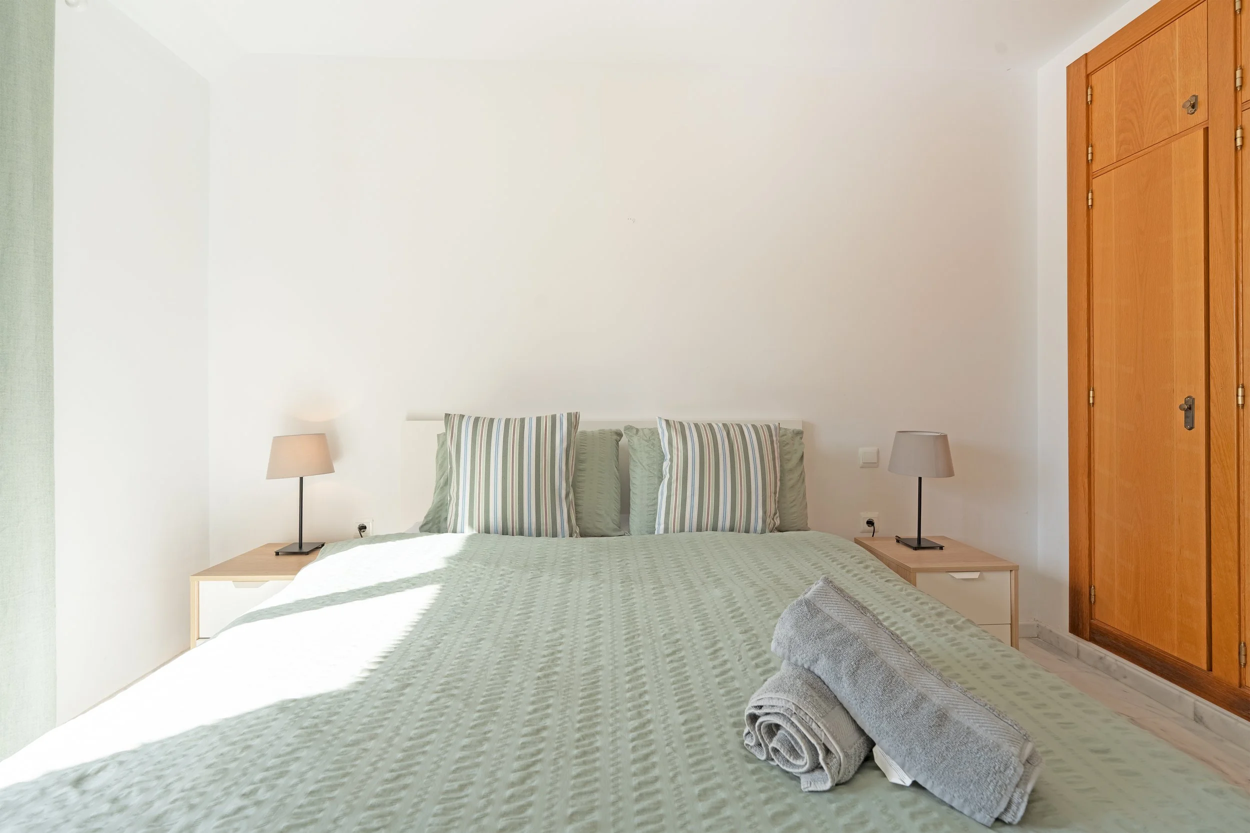 Bedroom with a large bed, three striped pillows, two bedside tables with lamps, a rolled towel near the bed, a wooden cupboard, and white walls.