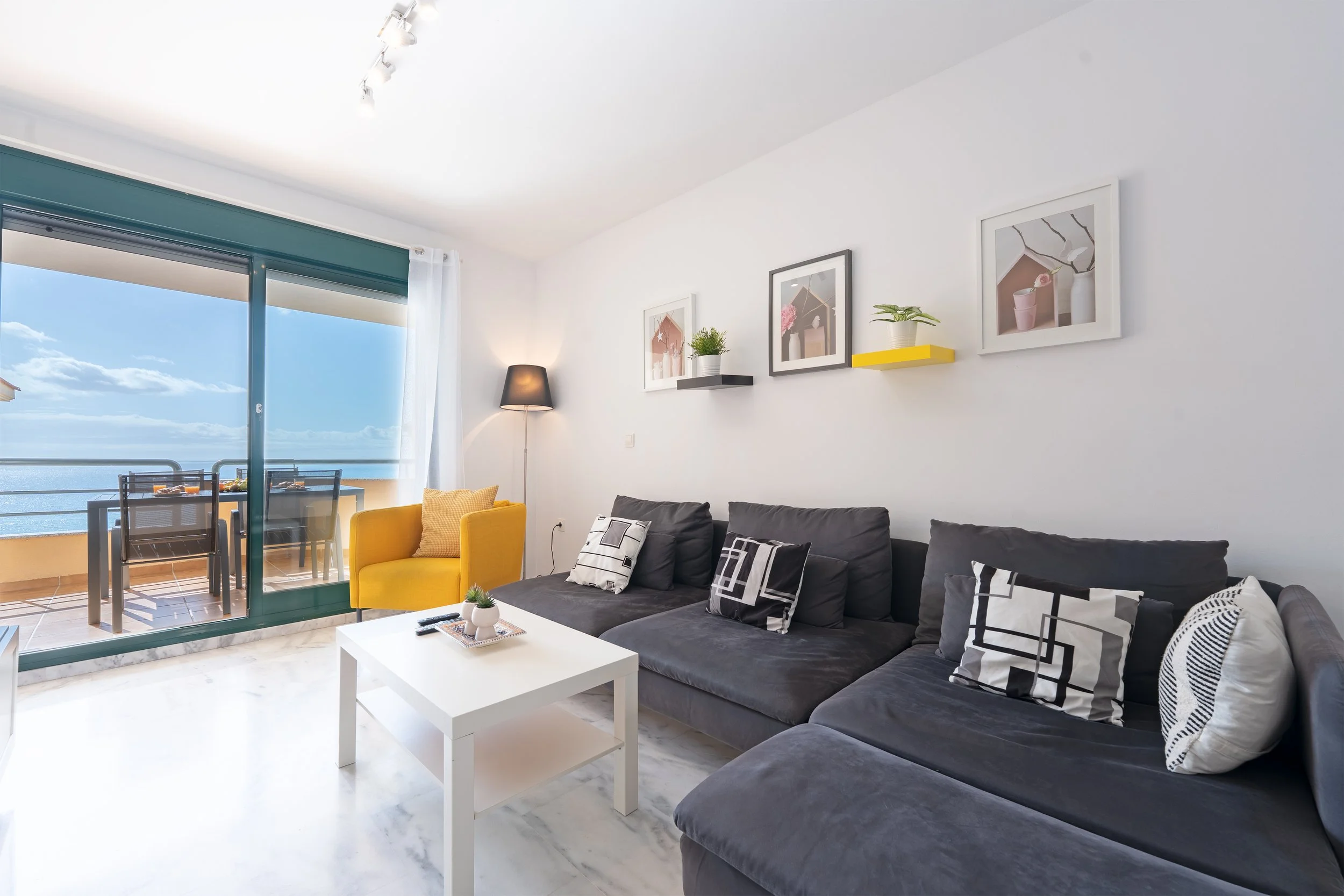Living room with a gray sofa, black and white pillows, white coffee table, yellow armchair, wall art, floor lamp, and balcony view of the ocean.