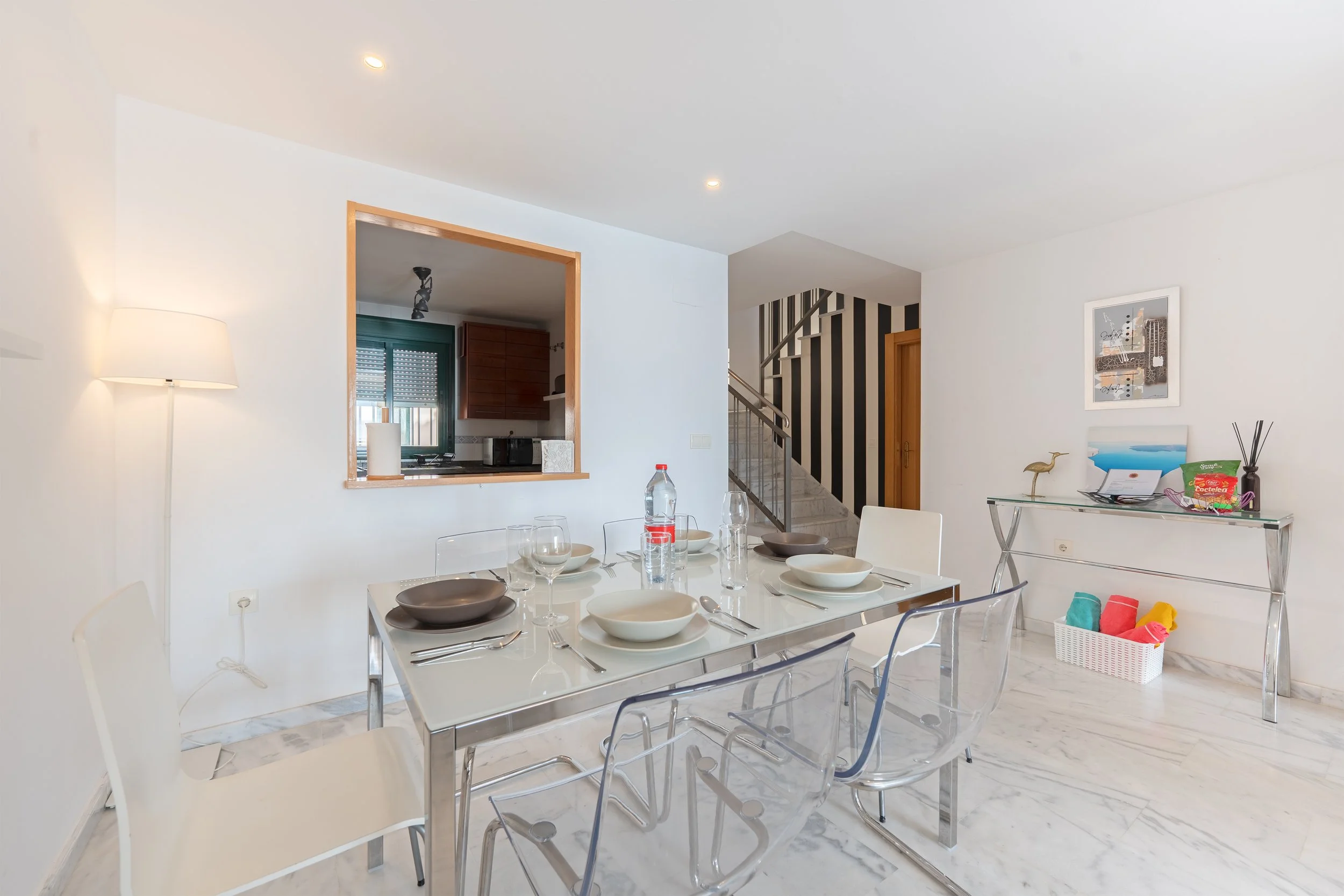 Dining room with glass table set for five, featuring bowls, plates, wine glasses, and a water bottle. Transparent and white chairs surround the table. The room has white walls, marble flooring, and a small window with a view into the kitchen. A conso