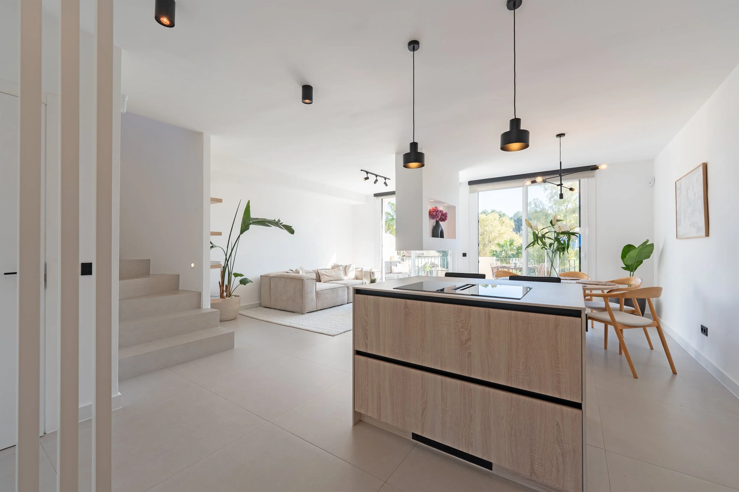 Bright, modern open-concept living and dining area with a kitchen island, beige sofas, wooden dining table and chairs, large windows, and indoor plants.