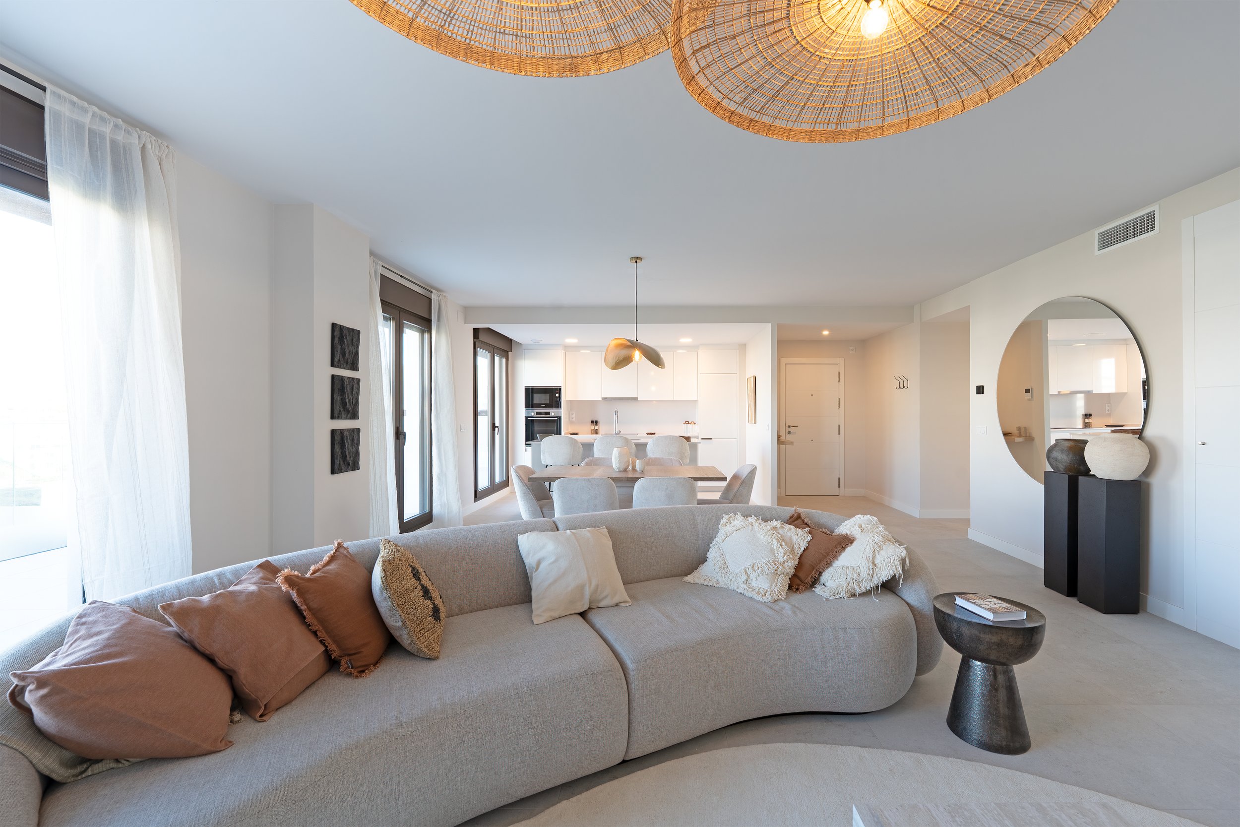 Modern, open-concept living room and dining area with neutral tones, a large beige sectional sofa with decorative pillows, round dining table with eight chairs, and a kitchen with white cabinetry in the background. Bright natural light coming through