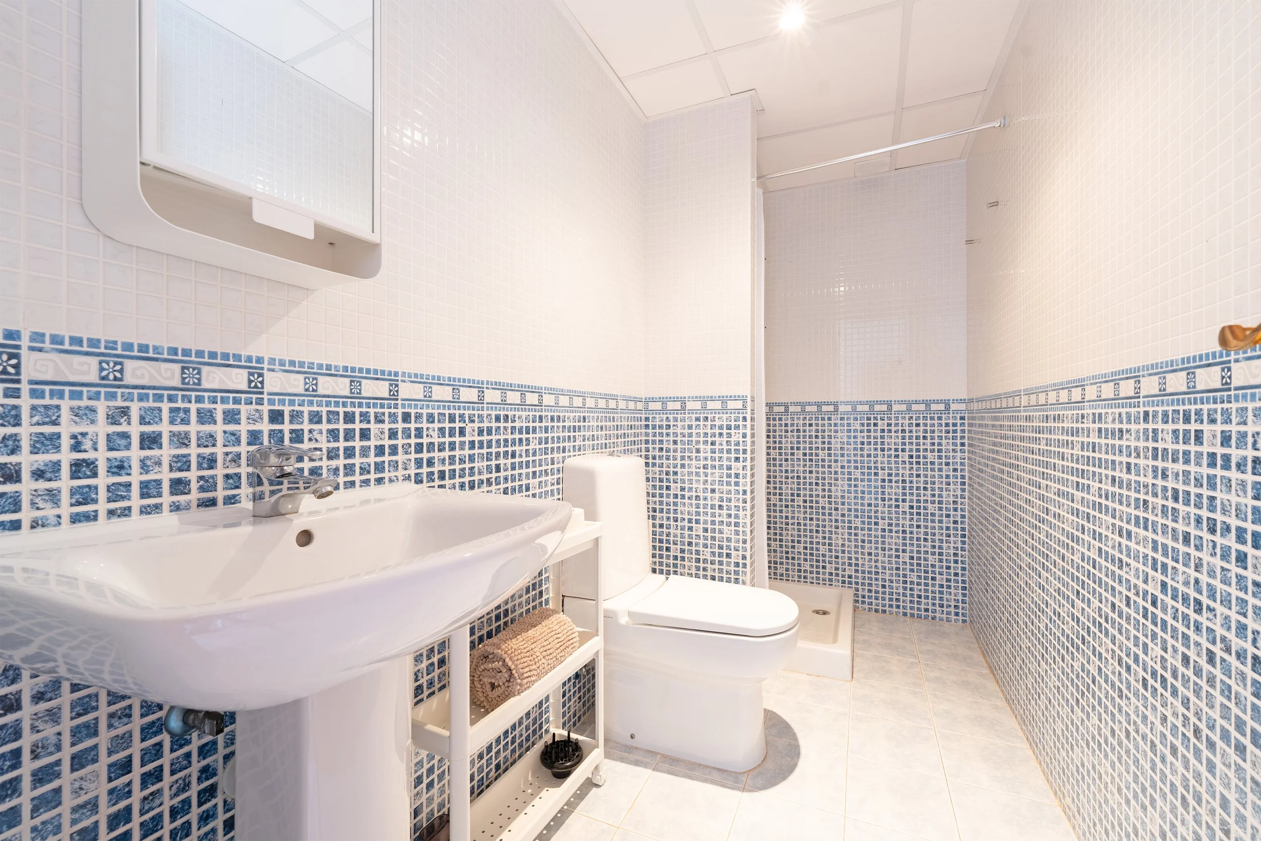 Small bathroom with white and blue mosaic tiles, a white sink, a toilet, a mirror cabinet, and a shower area with a curtain rod.