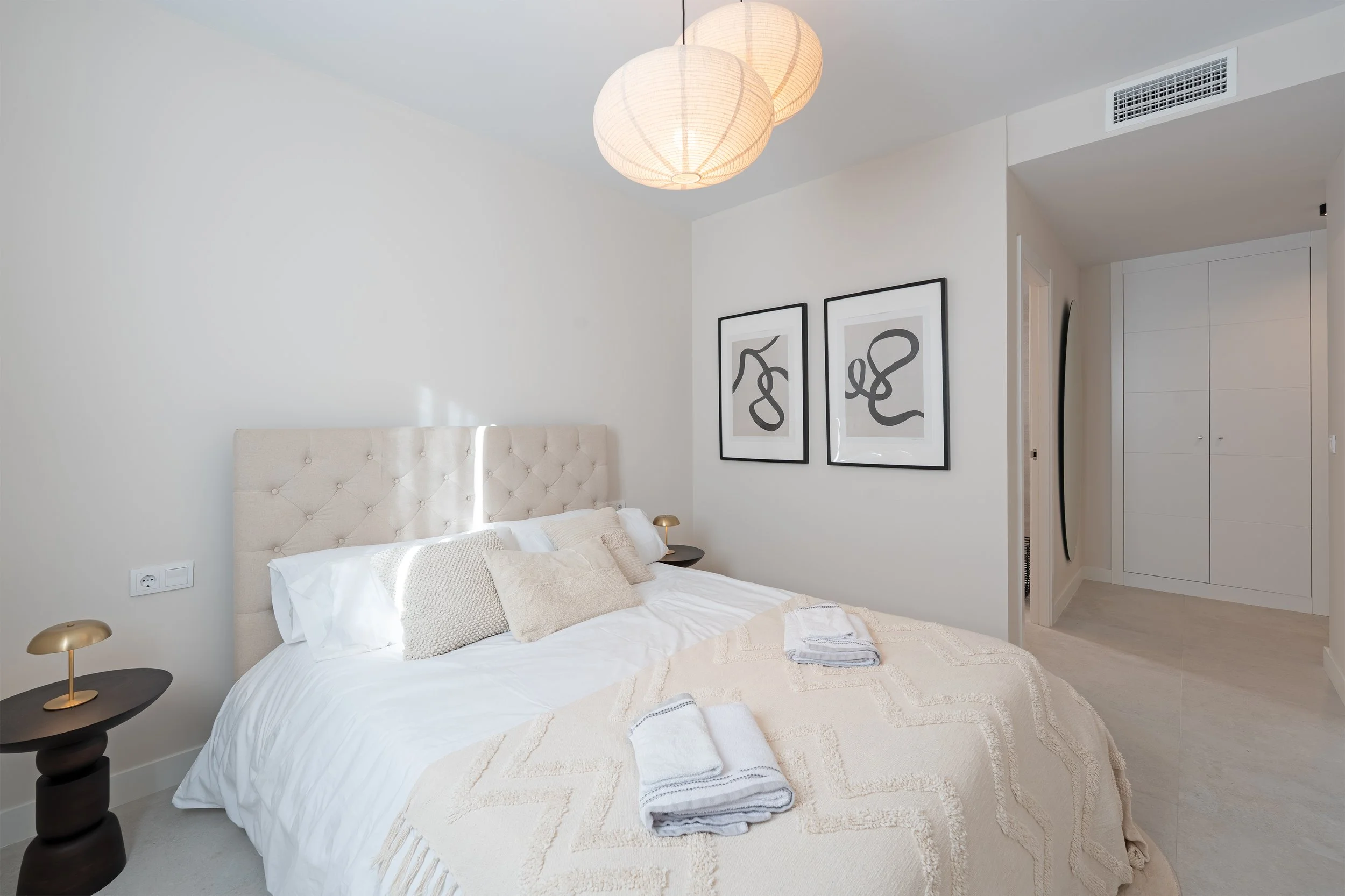 Bright bedroom with white bed, beige tufted headboard, folded towels on the bed, two bedside tables with golden lamps, and two abstract black-and-white framed artworks on the wall.
