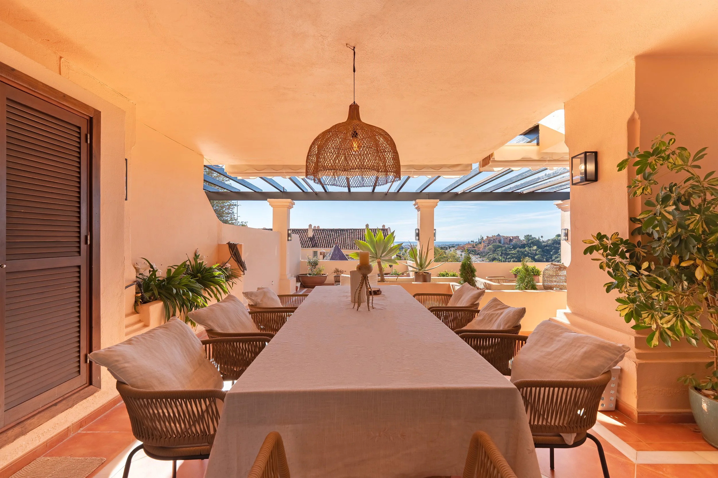 Outdoor dining area with a long table, surrounded by cushioned chairs, plants, and a view of a hillside town in the distance.
