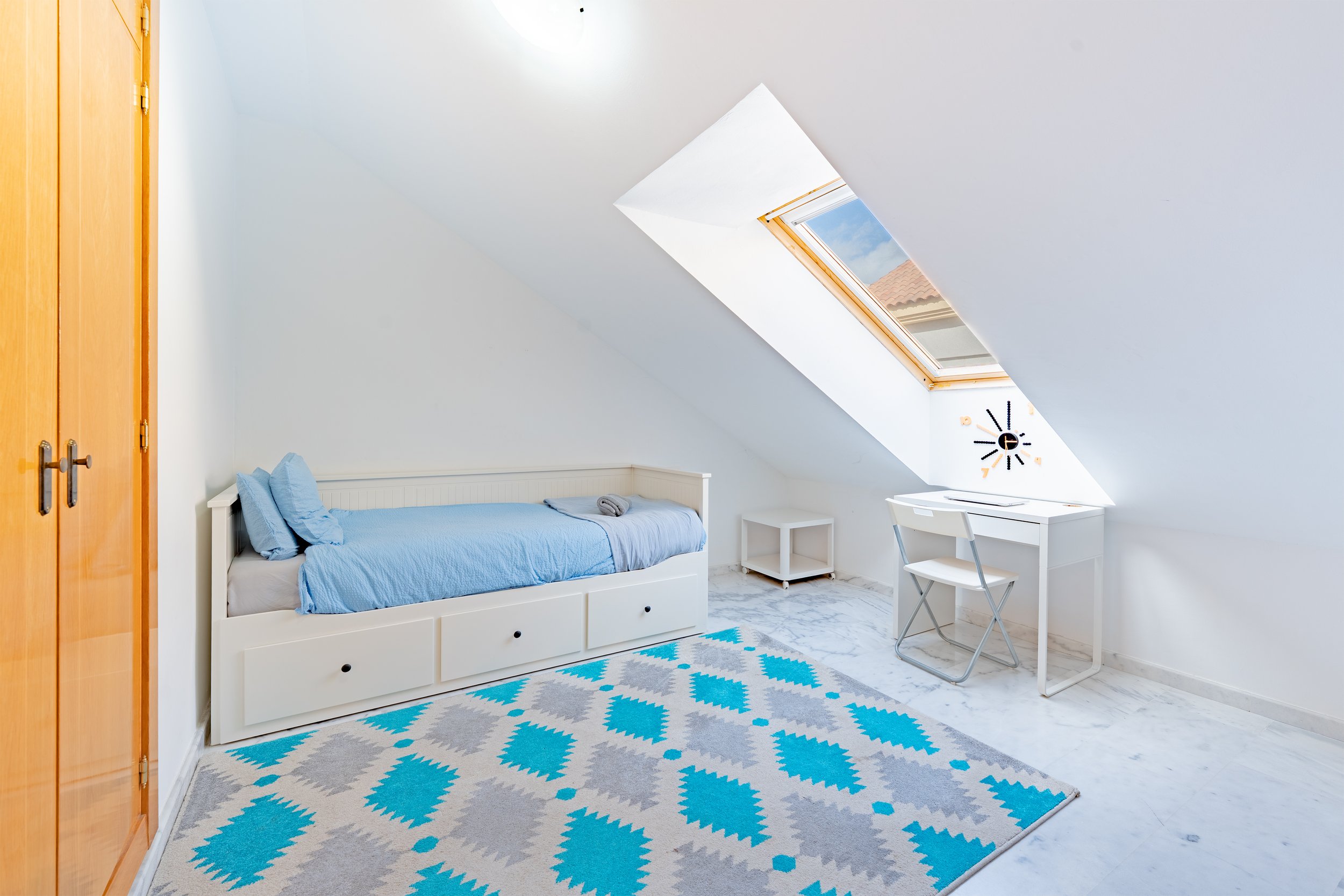 A bedroom with a sloped ceiling, a white bed with blue bedding, a window in the ceiling, a small white desk and chair, an orange wardrobe, a colorful blue and gray patterned rug, and a small white side table.