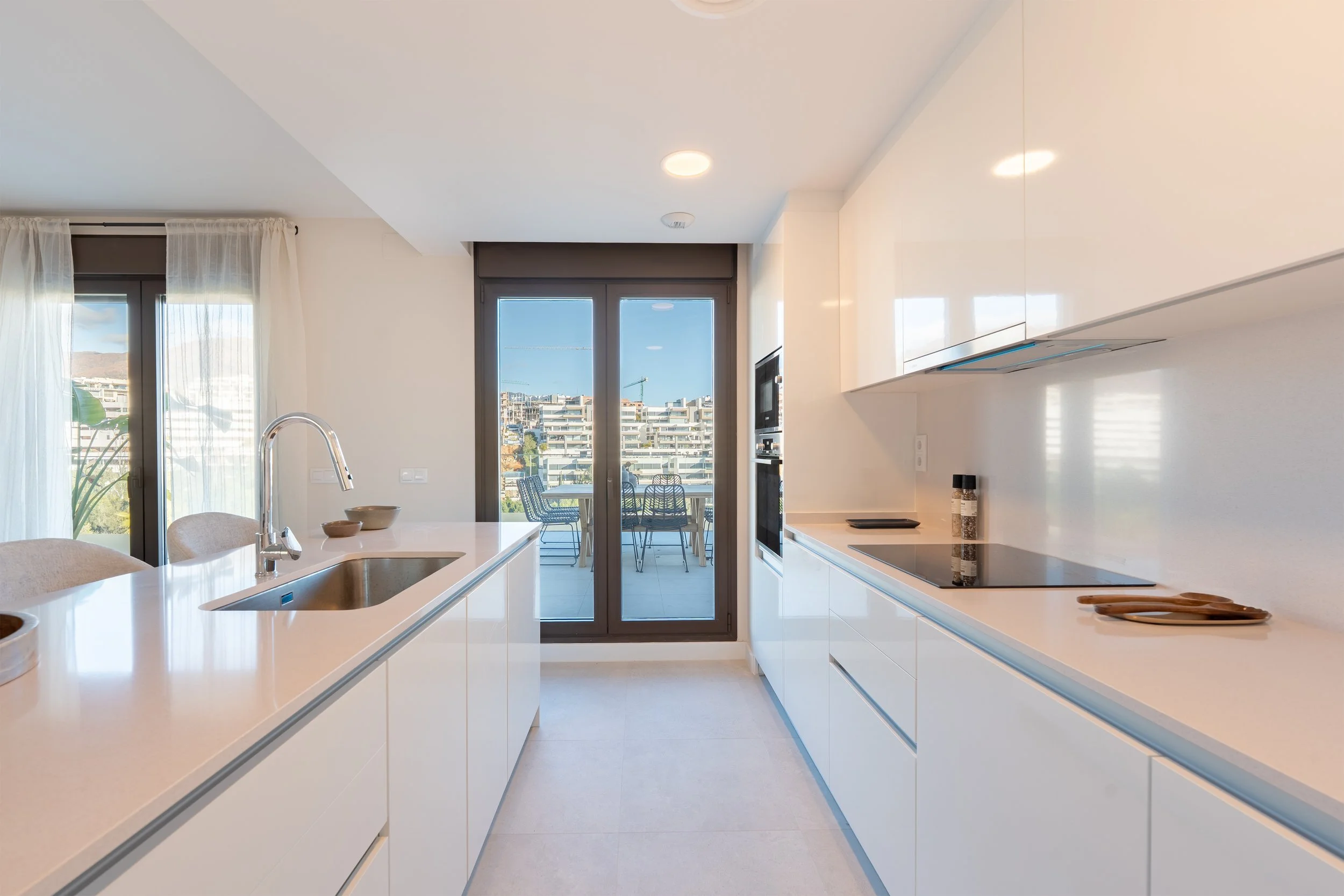 Modern white kitchen with a view to a balcony or terrace with outdoor furniture, large glass doors, minimalist design, and cityscape in the background.