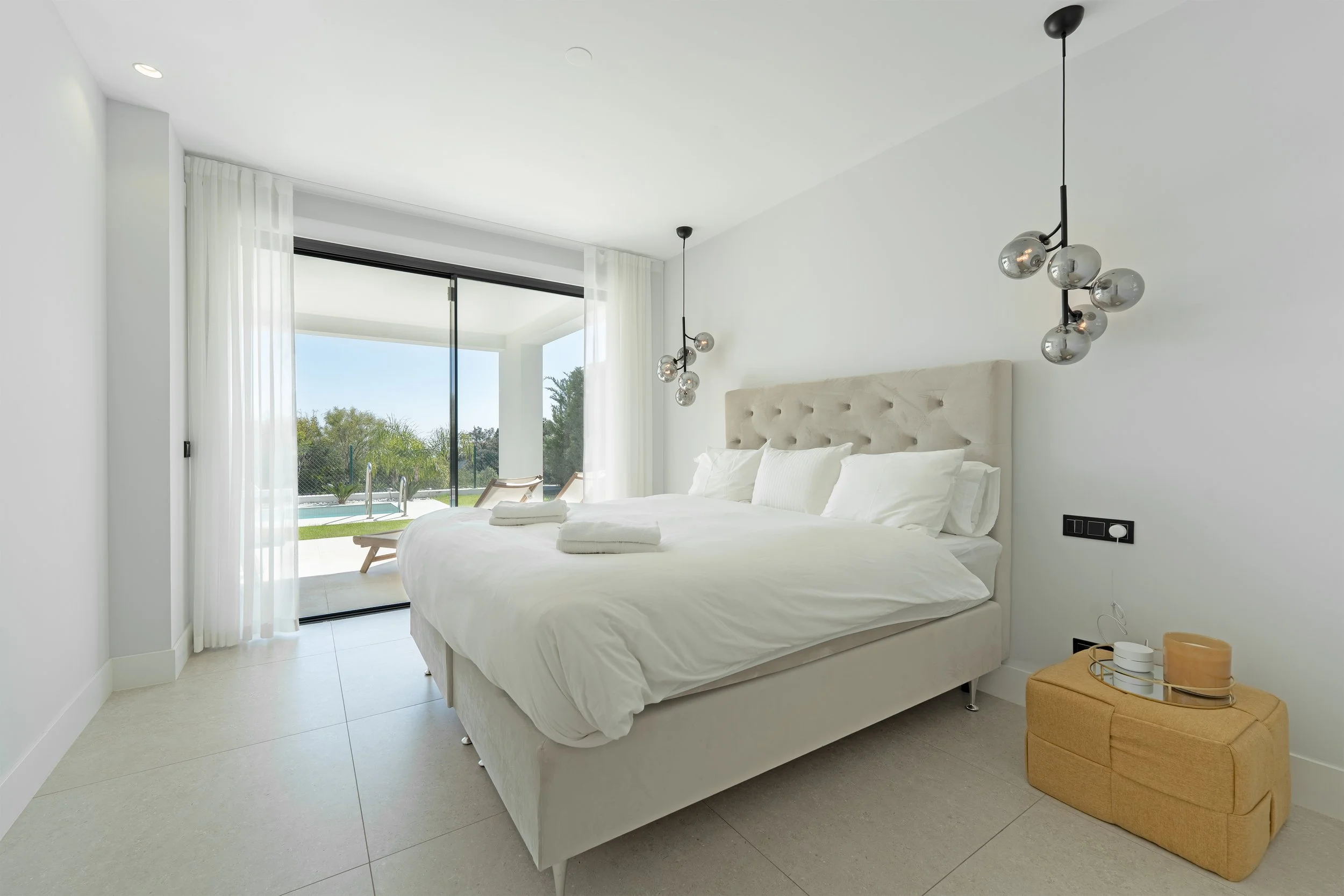 Modern bedroom with a large bed, white bedding, and a tufted headboard, next to sliding glass doors leading to an outdoor pool area.