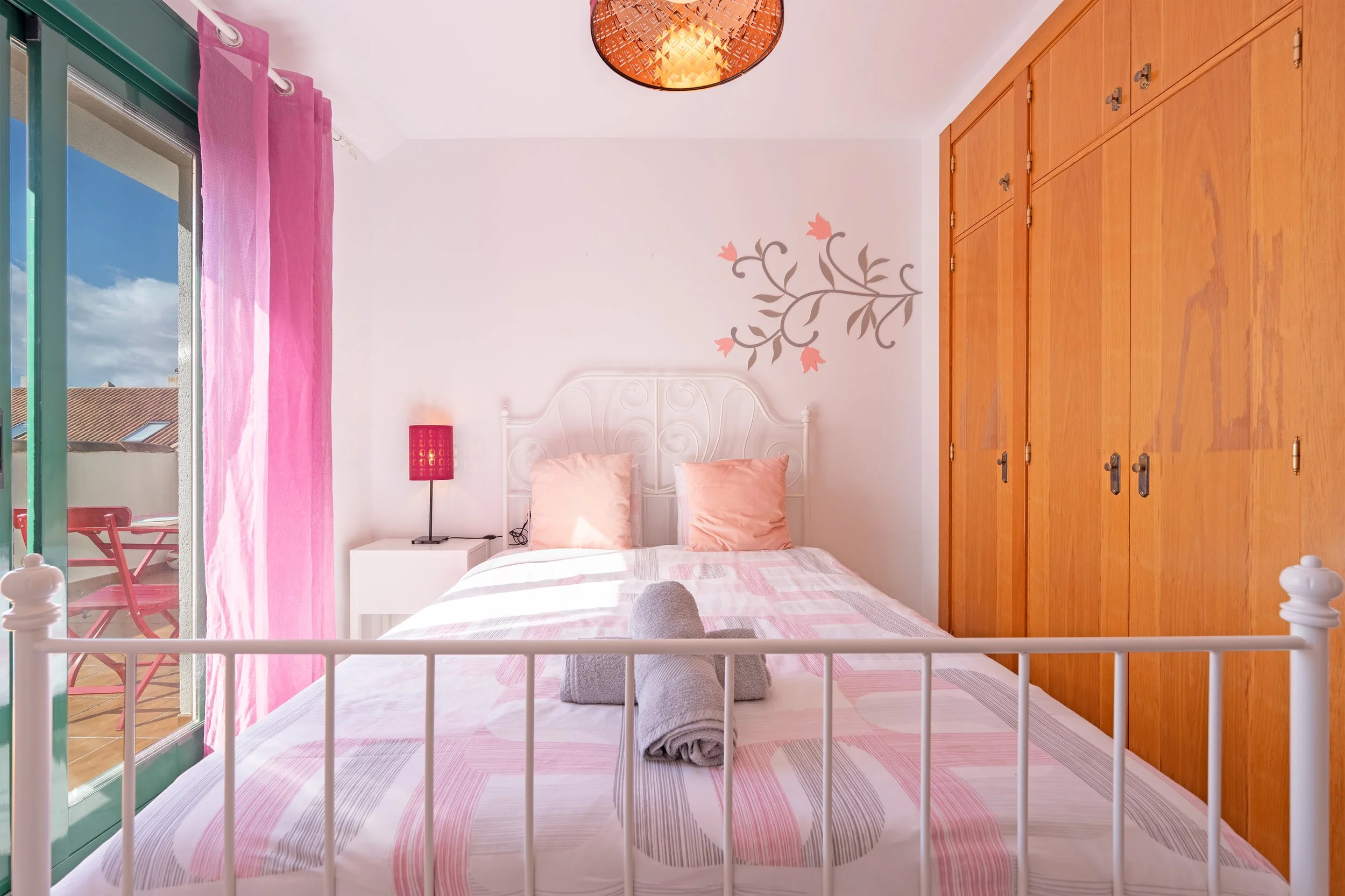Bright bedroom with white metal bed, pink pillows, folded gray towels on bed, white nightstand with pink lamp, pink curtains, glass door leading to balcony with outdoor furniture, wooden closet, floral wall decal, ceiling light.