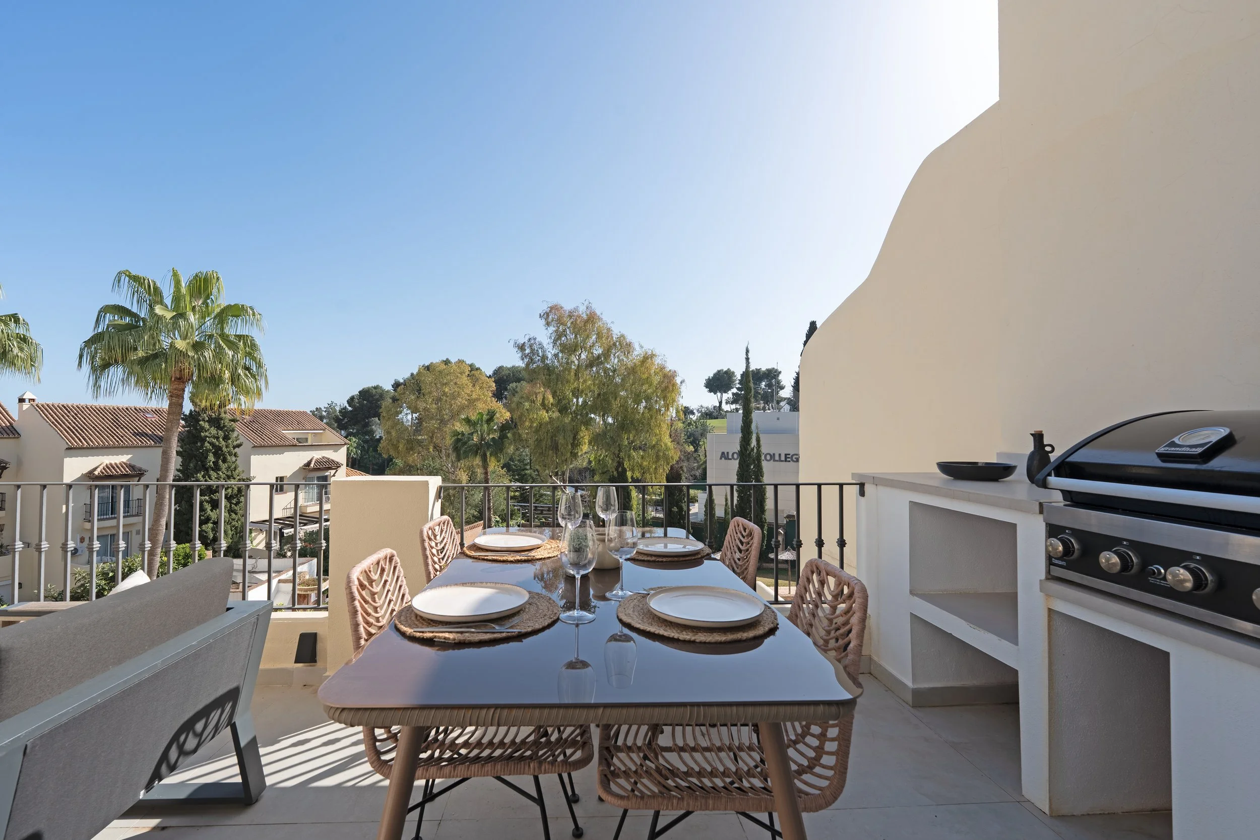 Outdoor balcony dining area with a wooden table set with plates, glasses, and napkins, surrounded by wicker chairs, with a barbecue grill on the right, overlooking a neighborhood with palm trees and a clear blue sky.