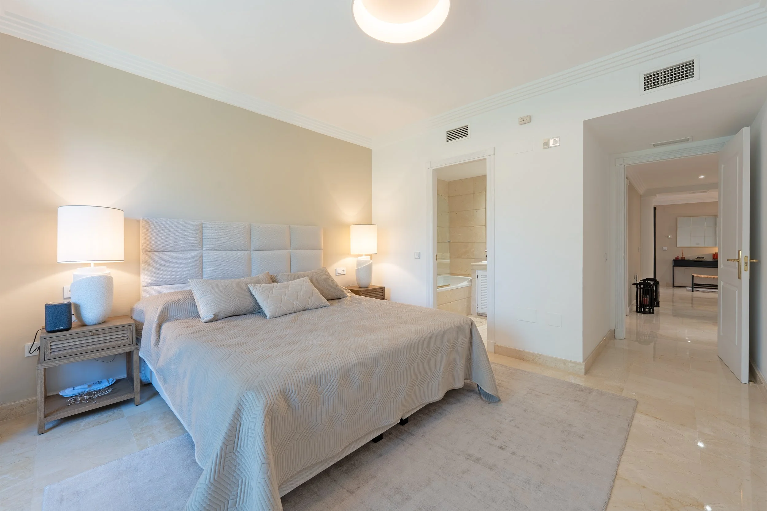 Modern bedroom with beige bedding, two nightstands with lamps, beige tiled floor, and open door leading to a hallway with additional rooms.
