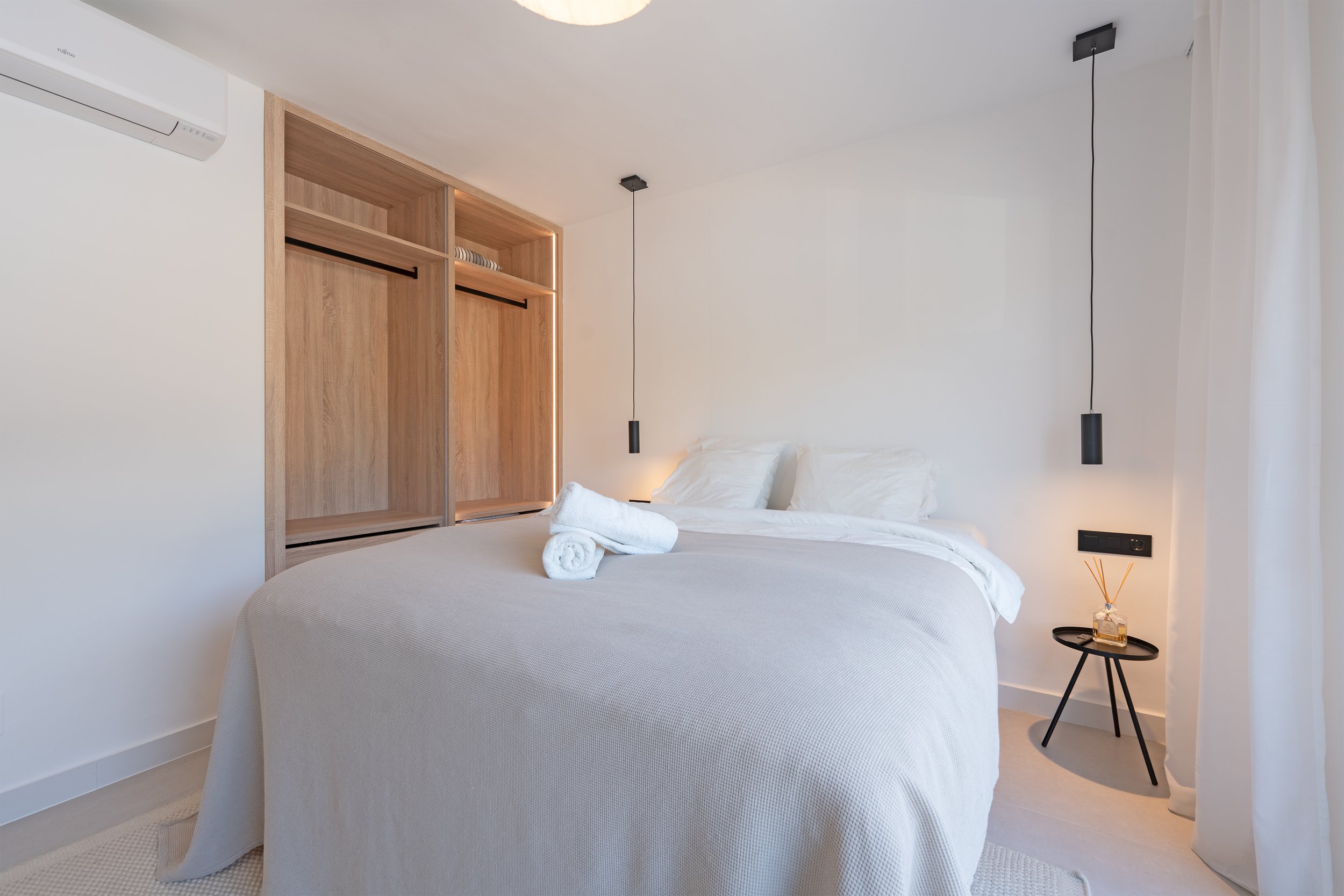 Minimalist bedroom with white bedding, a small black side table with a reed diffuser, white curtains, and a wooden wardrobe with open shelves and hanging space.