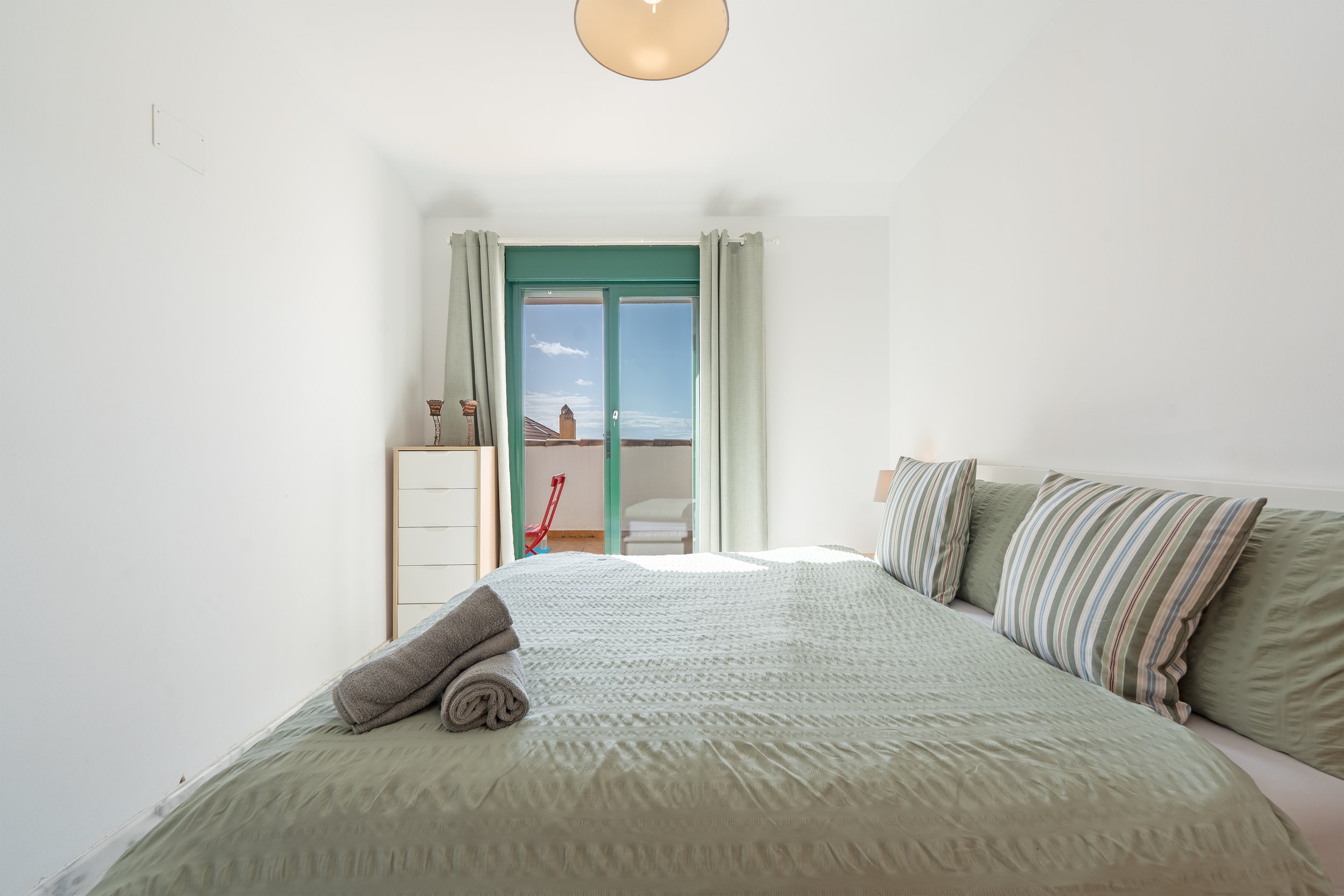 Bedroom with a bed, pillowcases with striped patterns, rolled towels on the bed, a chest of drawers, curtains, and a sliding glass door leading to a balcony with a view of the sky and rooftops.