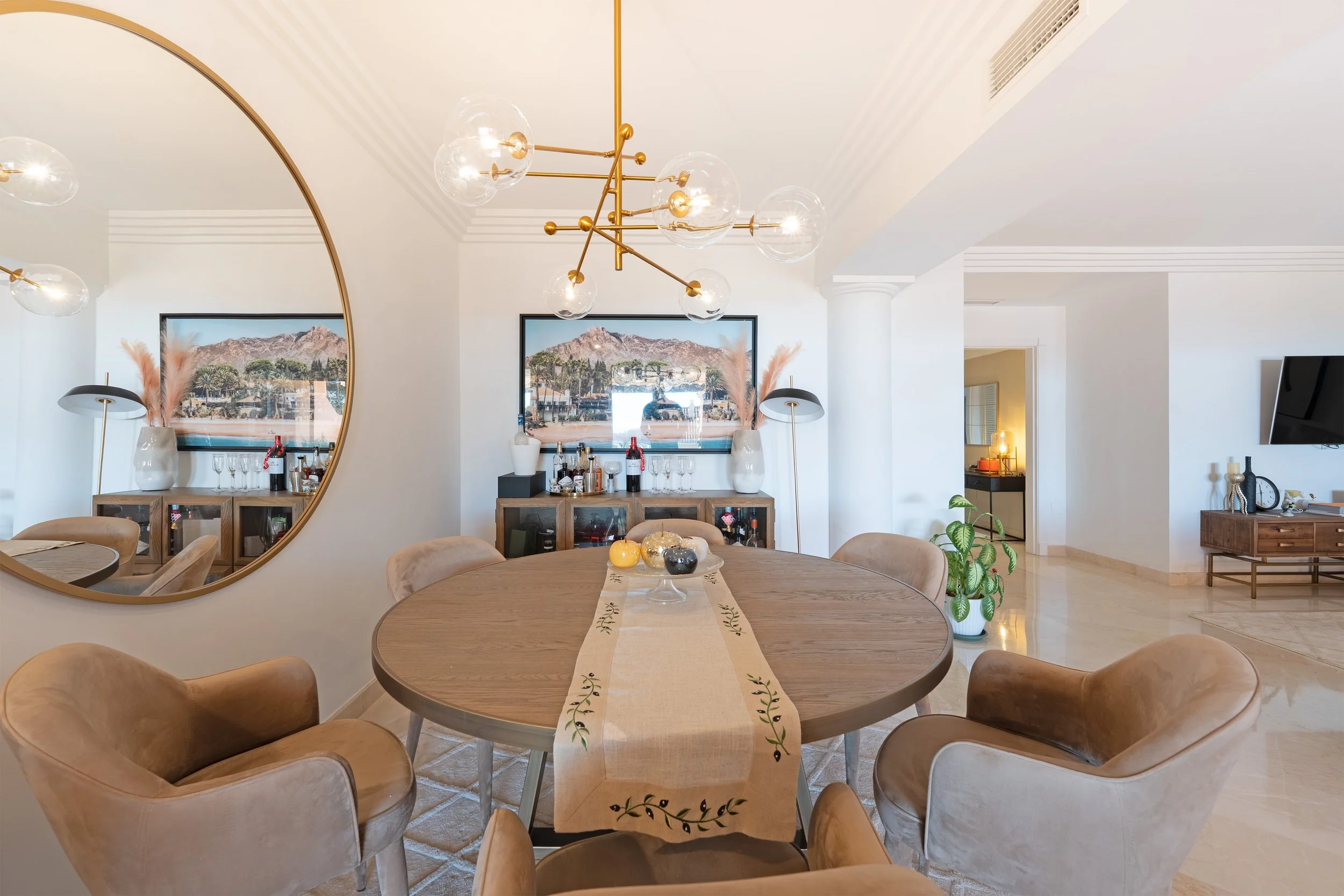 Modern dining room with a round wooden table, six beige upholstered chairs, and a long beige table runner with green embroidery. A chandelier with gold accents and multiple glass globes hangs above. A large round mirror on the left wall reflects part