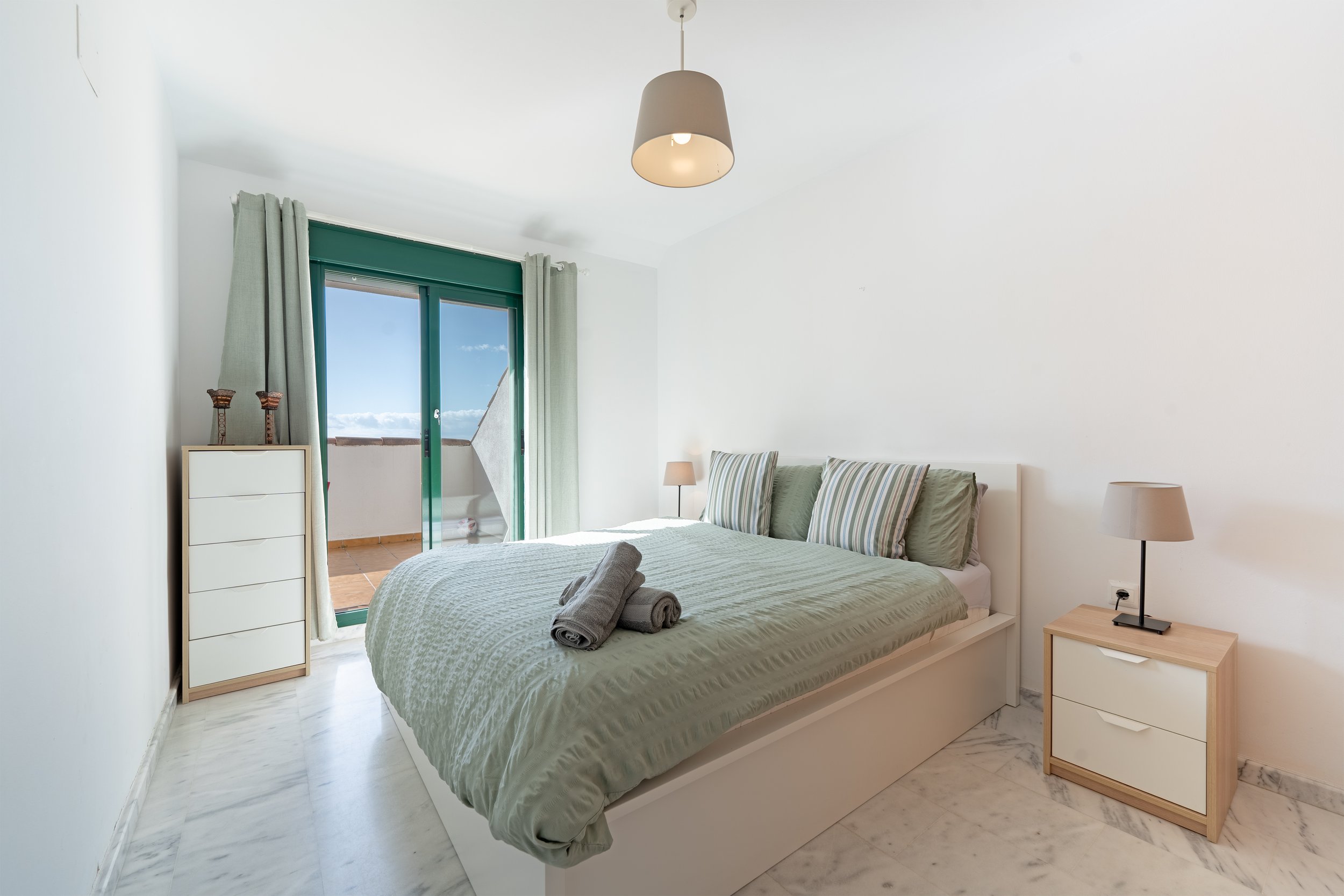 A bedroom with a bed, two nightstands, and a balcony door with green curtains. The bed has a light green duvet, four pillows with striped and solid covers, and two gray towels rolled on top. There is a lamp on each nightstand. The walls are white, an