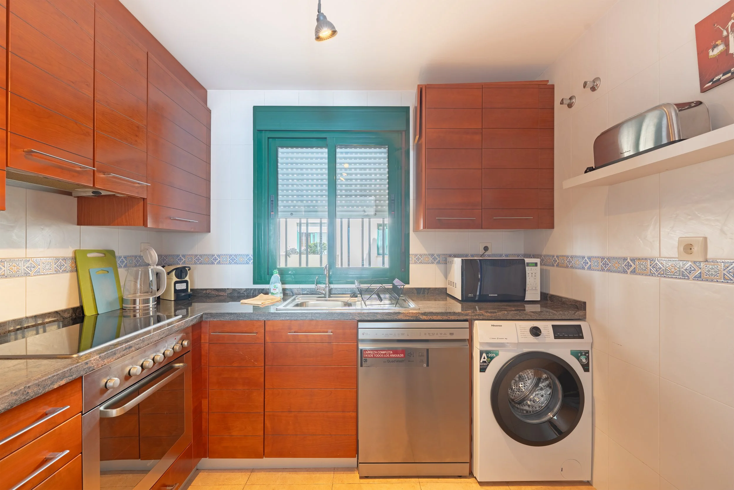Kitchen with wooden cabinets, green window, stainless steel appliances, and a washing machine. Items include a microwave, toaster, kettle, and cleaning supplies.