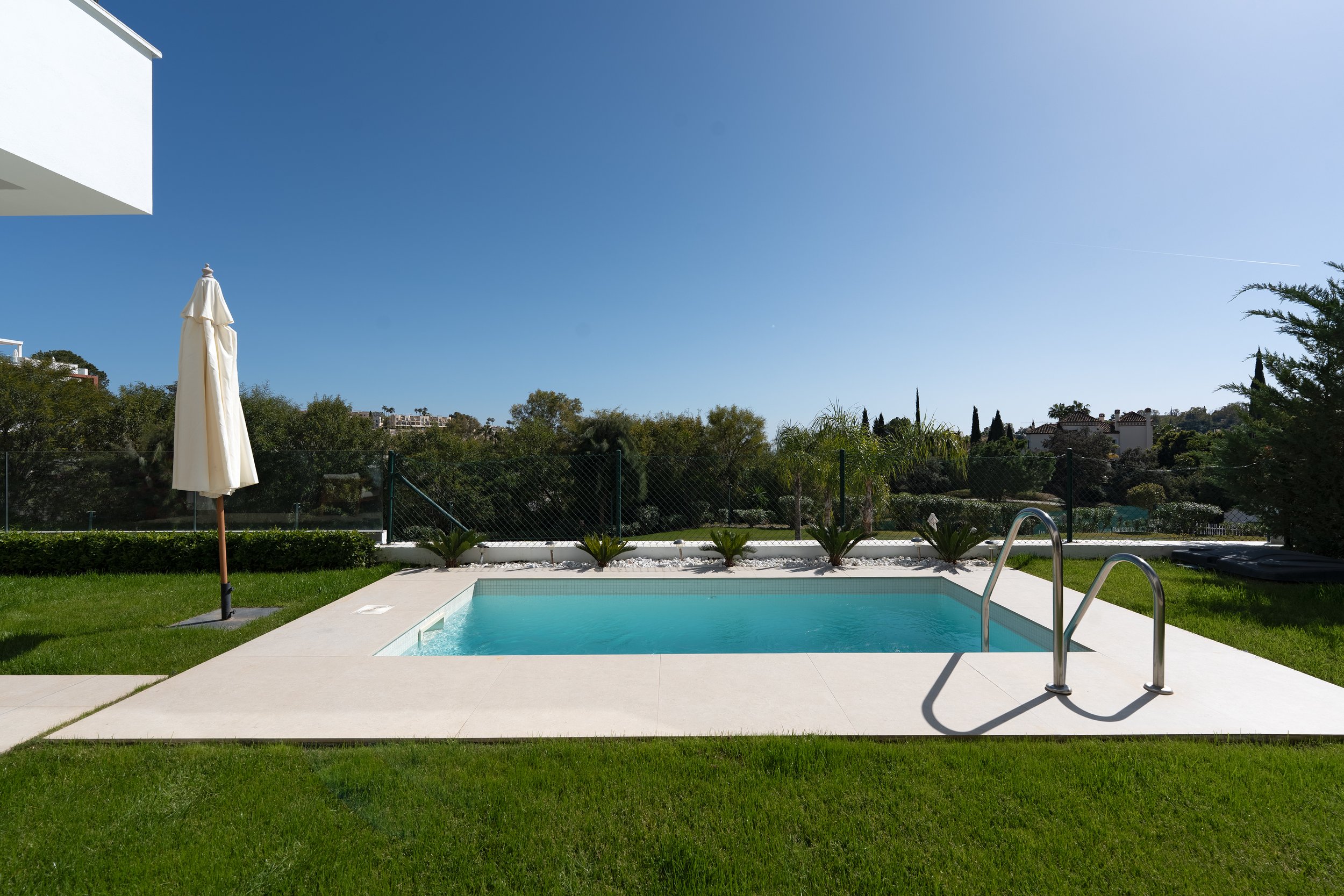 A backyard with a small swimming pool, a white patio, and an umbrella on a grassy area under a clear blue sky.