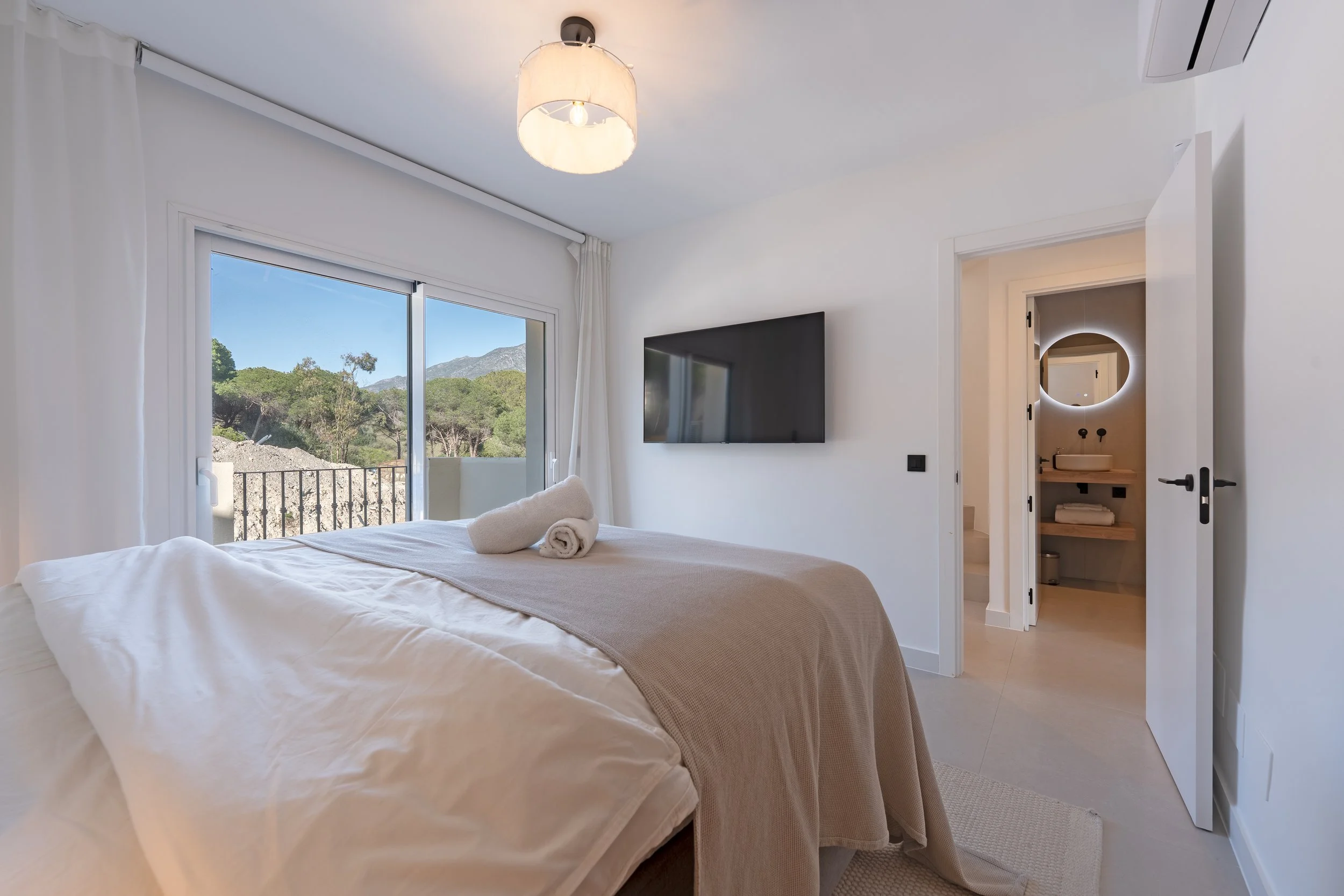 Bright bedroom with white walls, a large bed with white bedding and rolled towels on top, a sliding door with a view of trees and mountains, a wall-mounted TV, and an en-suite bathroom with a circular mirror and wooden shelves.