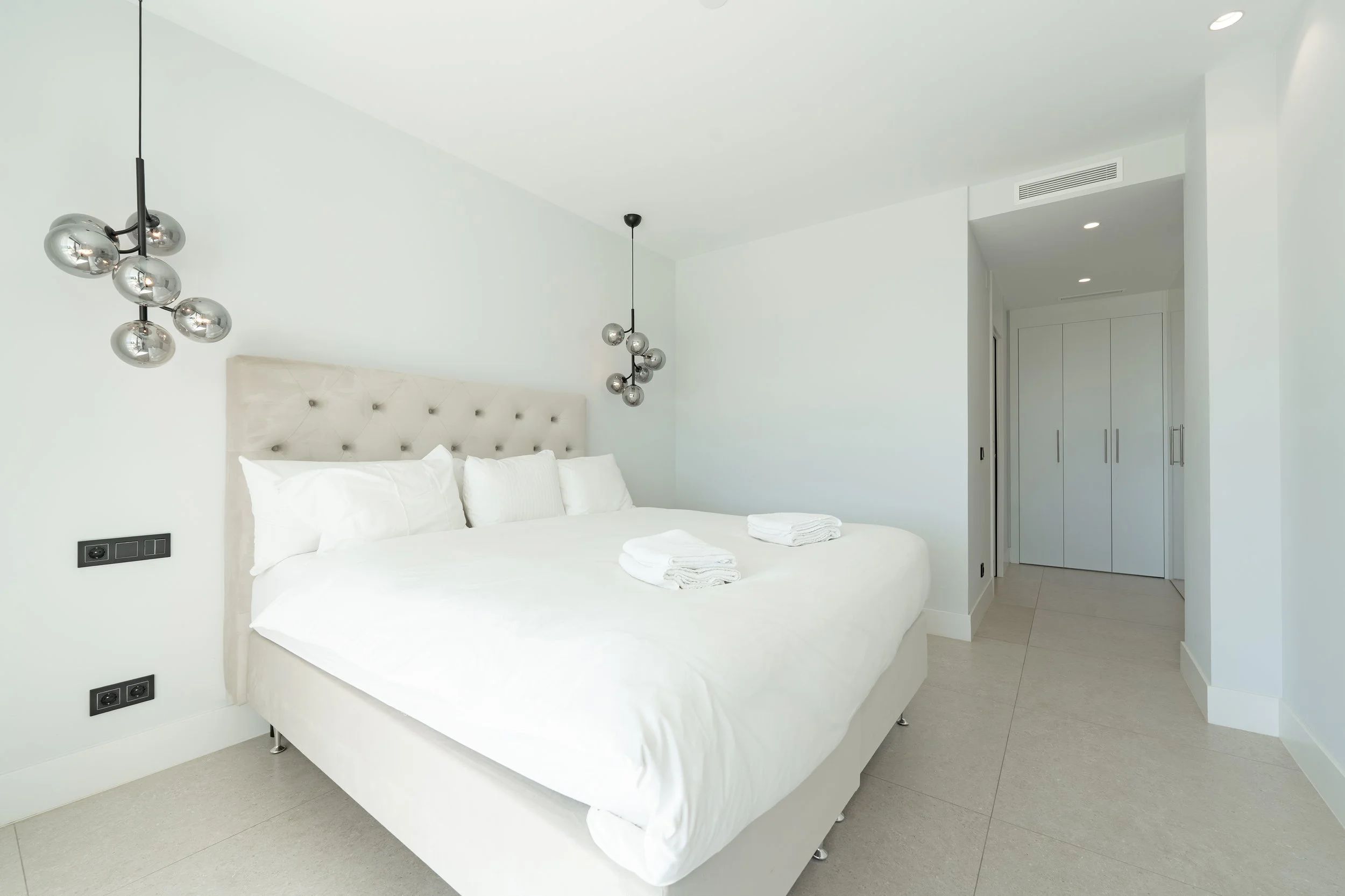 Minimalist bedroom with a white bed, plush headboard, white bedding, and two folded towels. Modern hanging silver globe light fixtures on either side of the bed. White walls, tiled floor, and a door to a closet in the background.