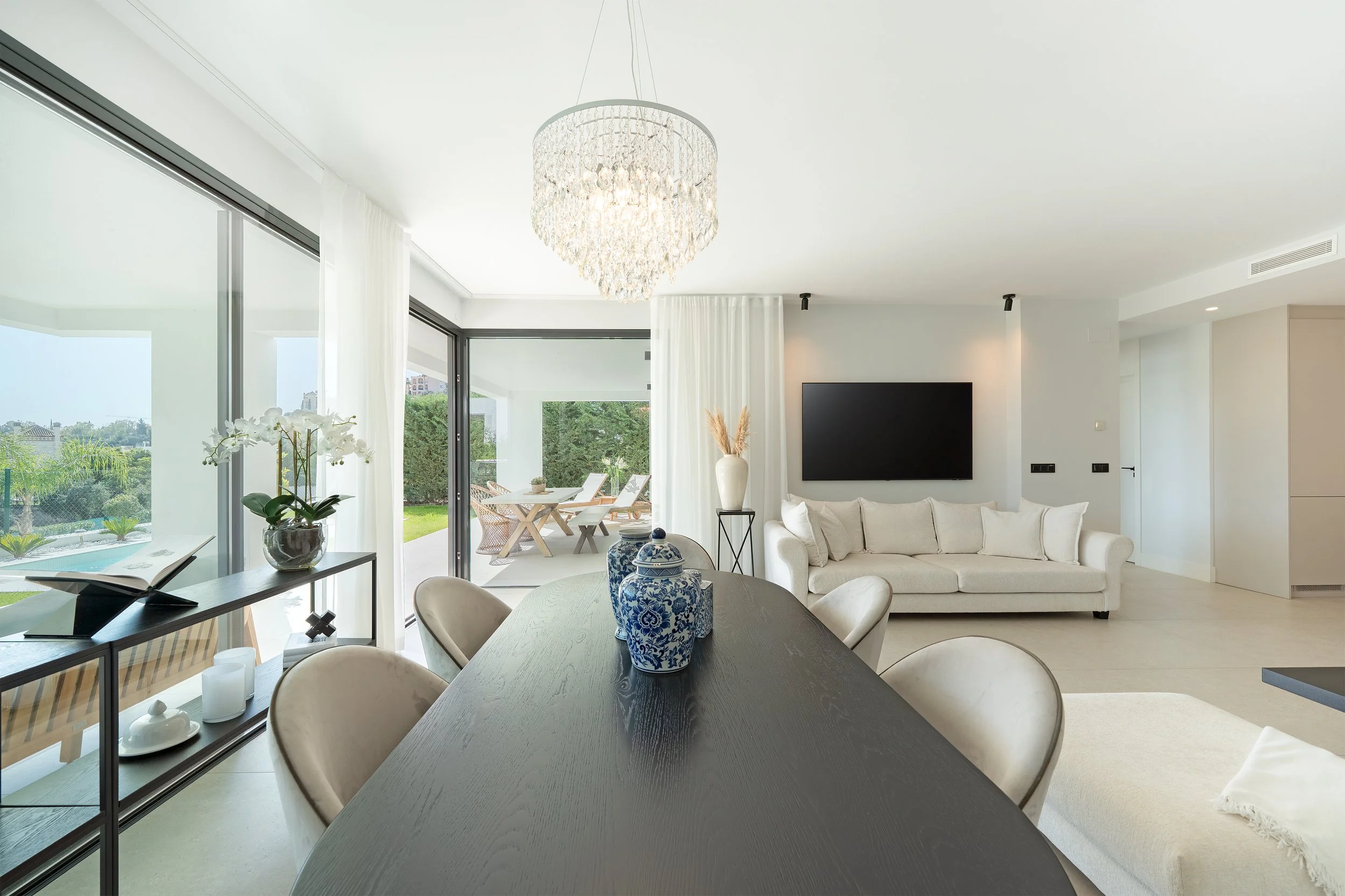 Bright modern living room with a dining table, beige chairs, a white sofa, decorative vases, and large windows showing outdoor patio and greenery.