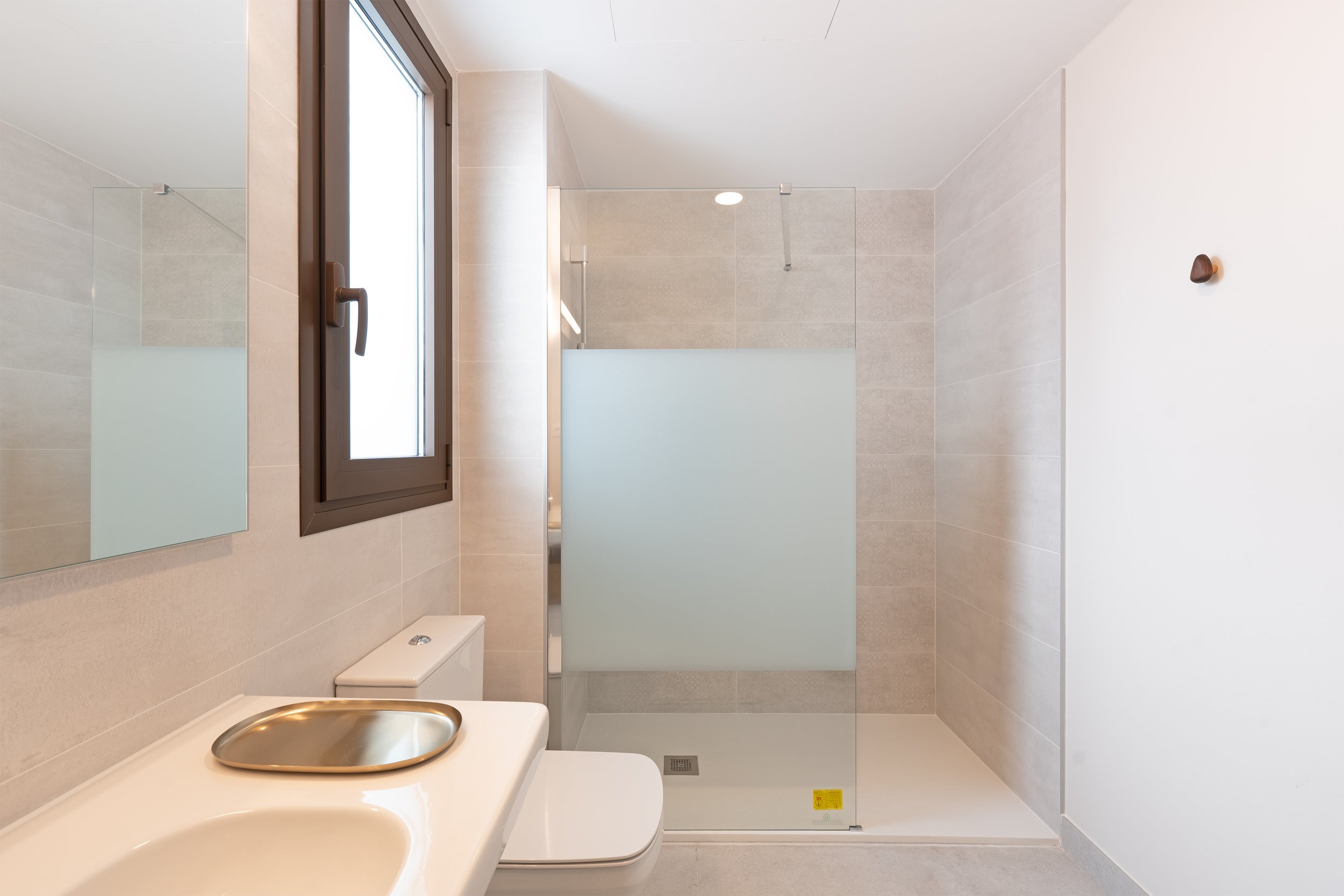 Modern bathroom with a frosted glass shower enclosure, a toilet, a small window, and a mirror on the beige tiled wall.