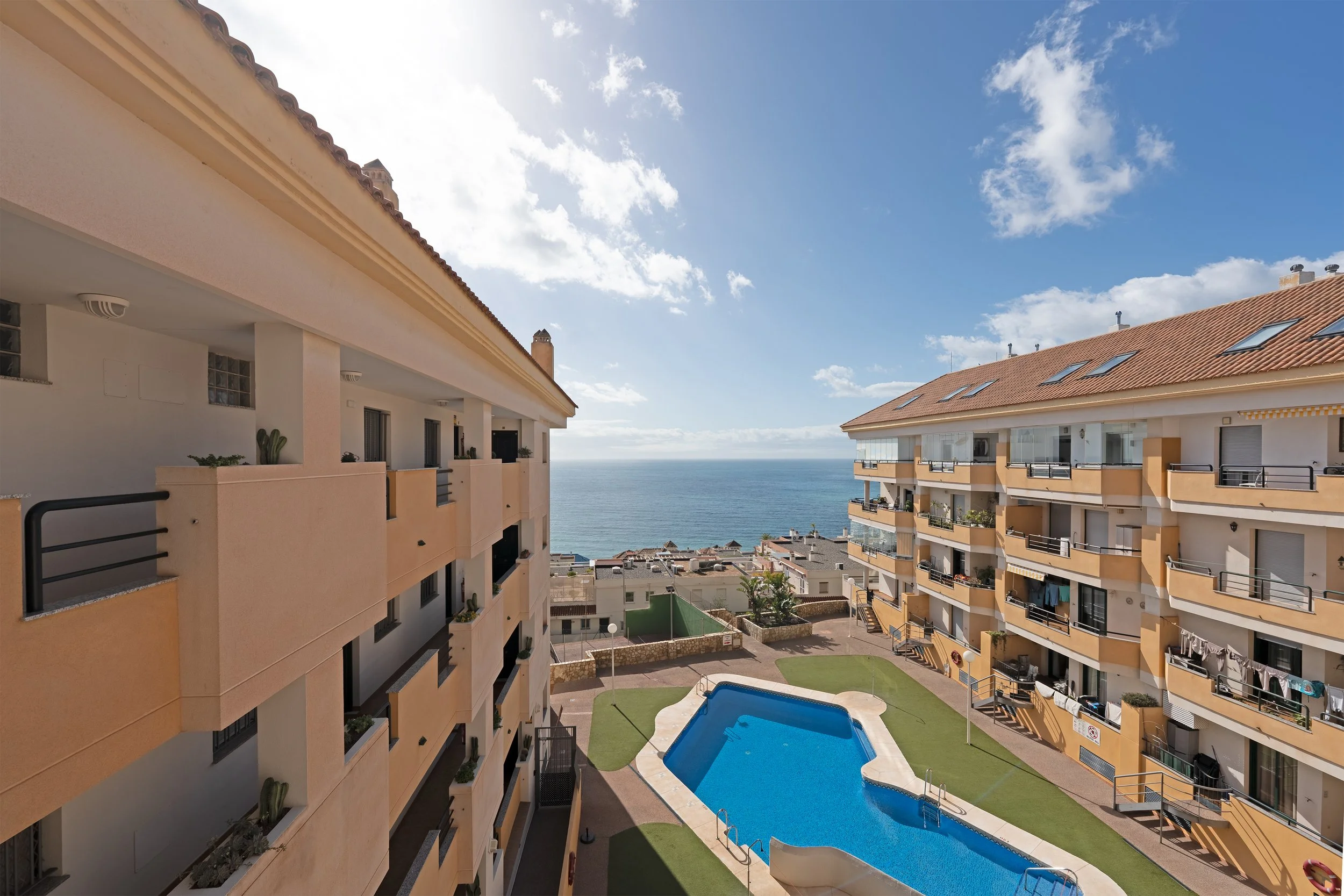 View of a hotel courtyard with a swimming pool, surrounded by balconies of two apartment buildings, overlooking the ocean under a partly cloudy sky.