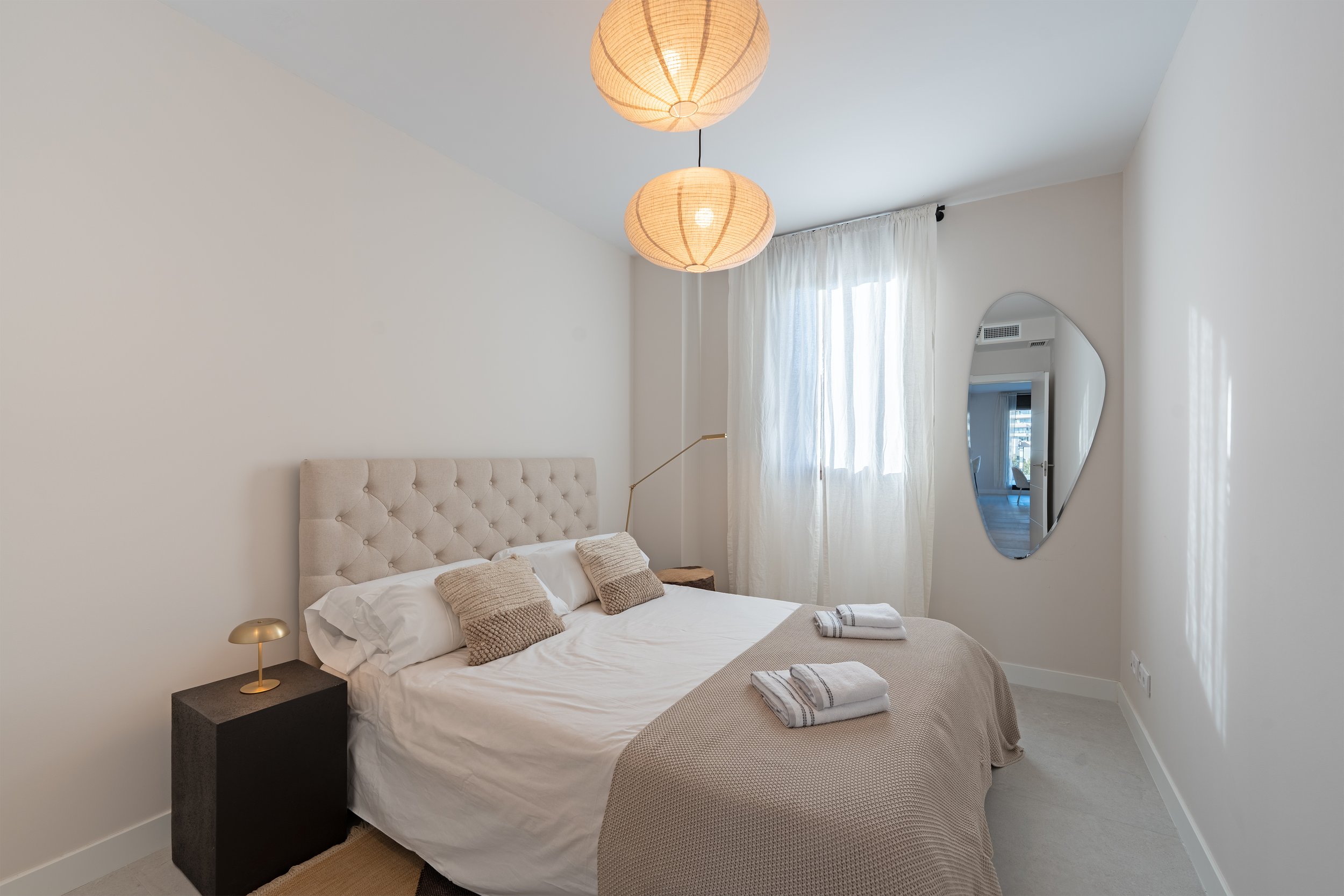 A neatly made bedroom with a tufted beige headboard, two beige pillows, and white sheets. There is a beige throw blanket at the foot of the bed with two folded towels on top. A black nightstand with a small gold lamp is beside the bed. The room has w