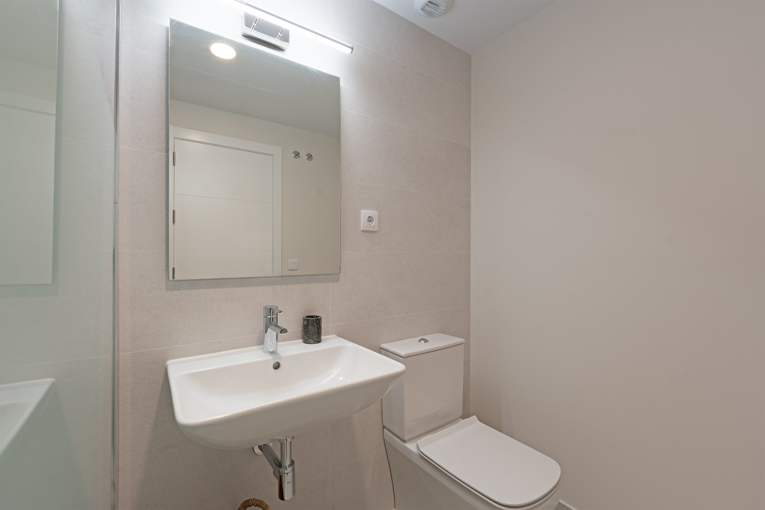 Small modern bathroom with a white wall-mounted sink, a rectangular mirror, and a white toilet, with beige tiled walls.