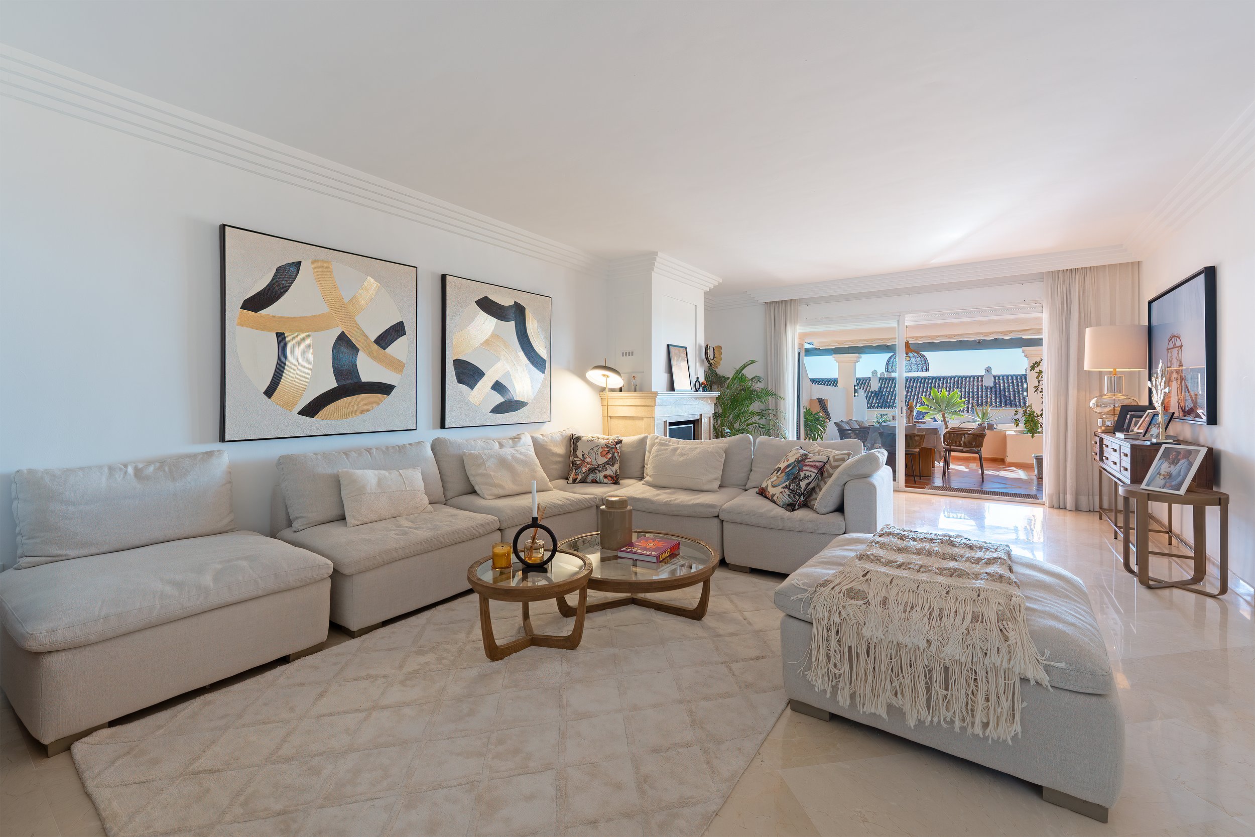Spacious living room with white sofa, colorful cushions, abstract art on the wall, and a balcony with outdoor furniture and plants.