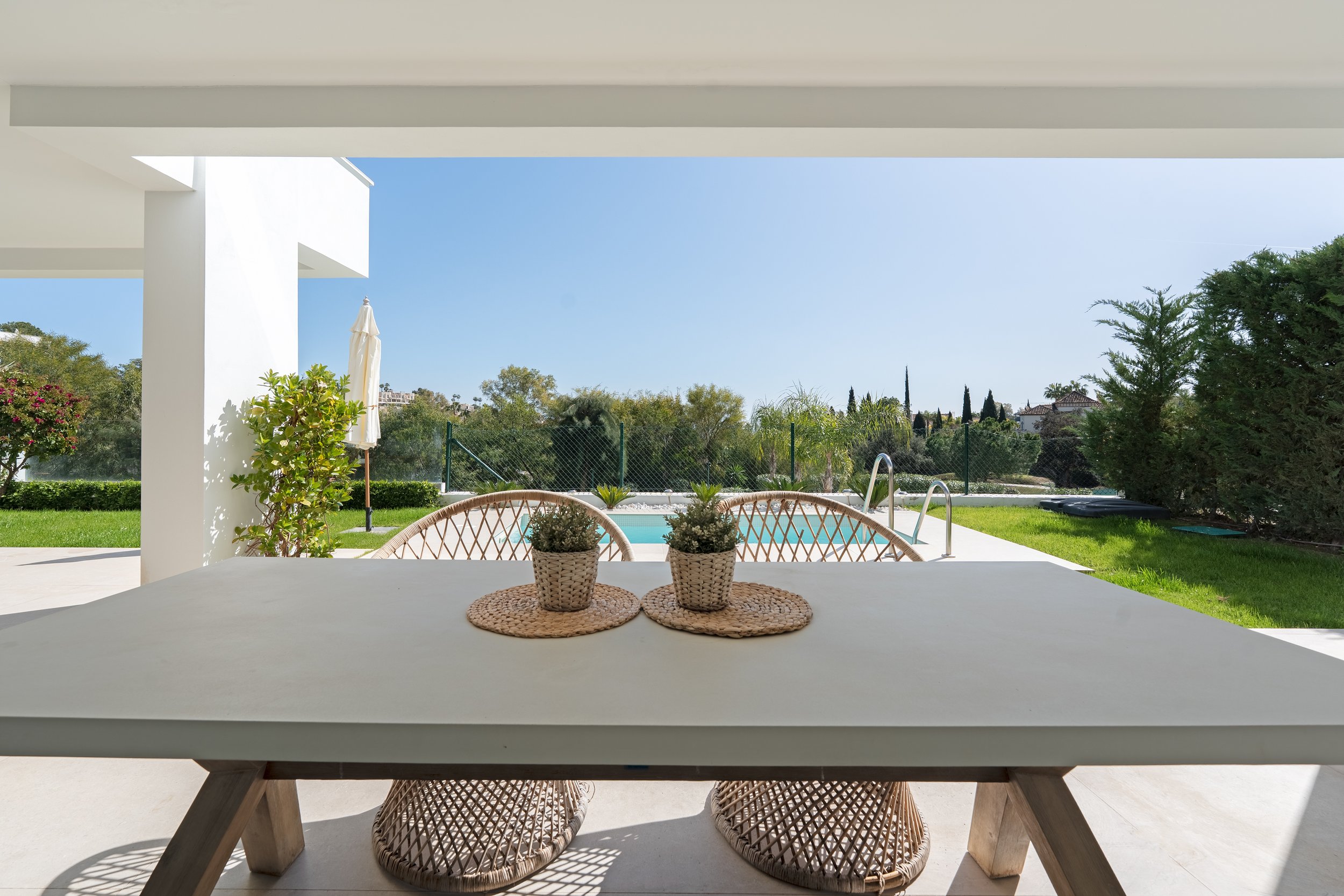 View of backyard patio with table, two wicker chairs, potted plants, green lawn, trees, and a pool under a clear blue sky.