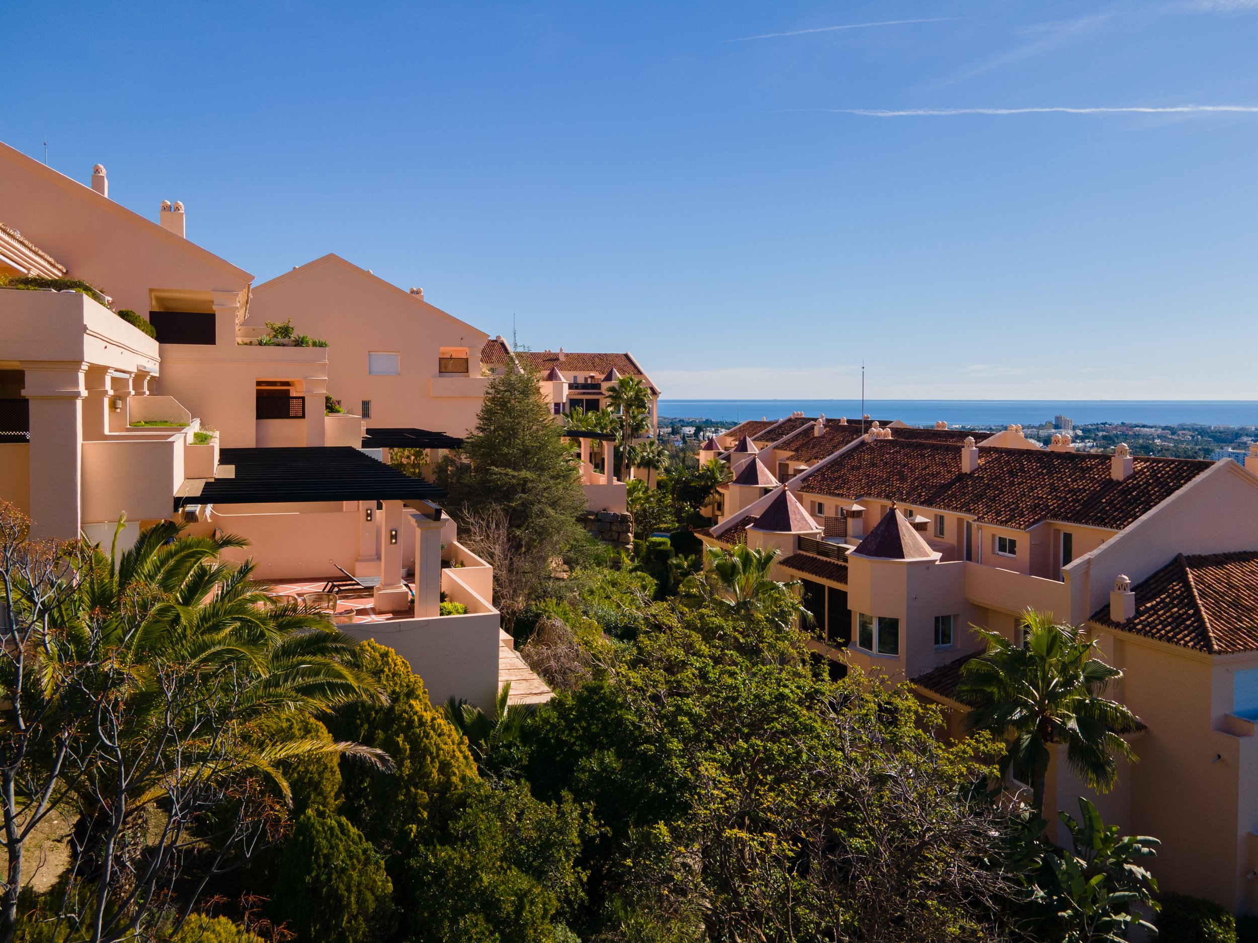 A view of residential houses with terracotta roofs and lush green trees, overlooking a coastal city and the ocean under a clear blue sky.