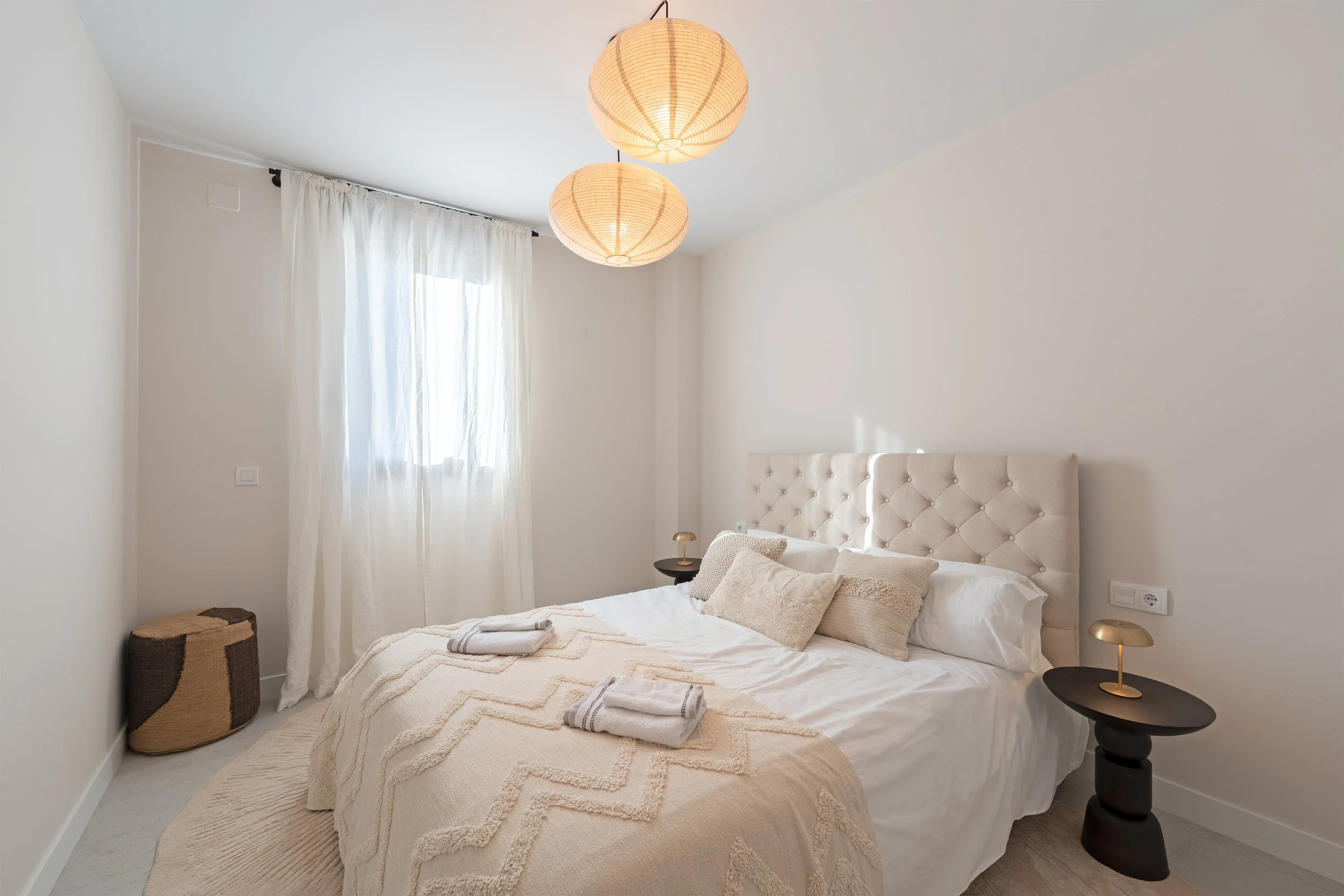 A minimalist bedroom with a white bed, beige headboard, and neutral decor. There are two black bedside tables with small golden lamps, white curtains on a window, and woven baskets in the corner. Two rolled towels are on the bed.