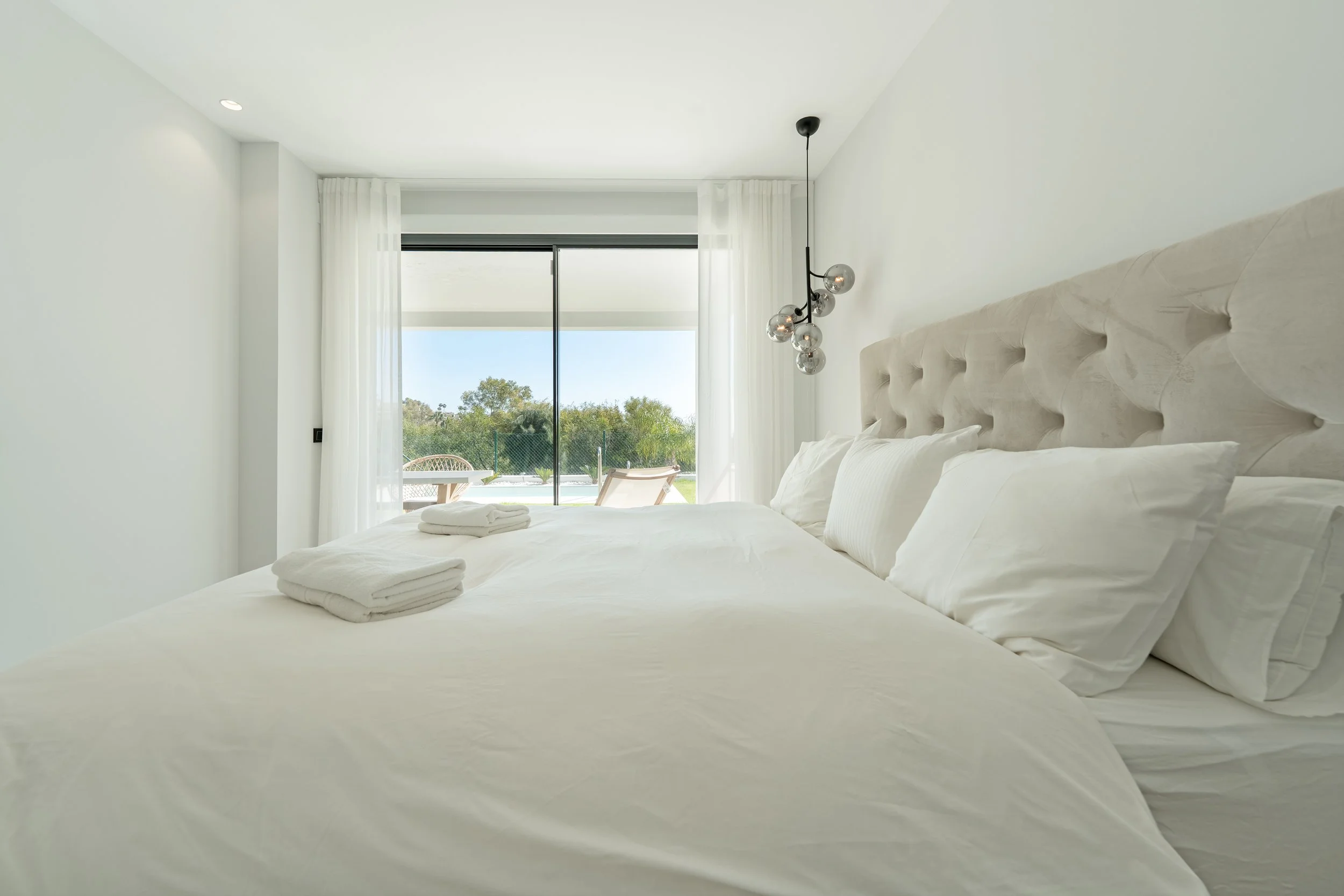 Bright modern bedroom with white bedding, a tufted beige headboard, and a sliding glass door leading to an outdoor area with chairs and trees.