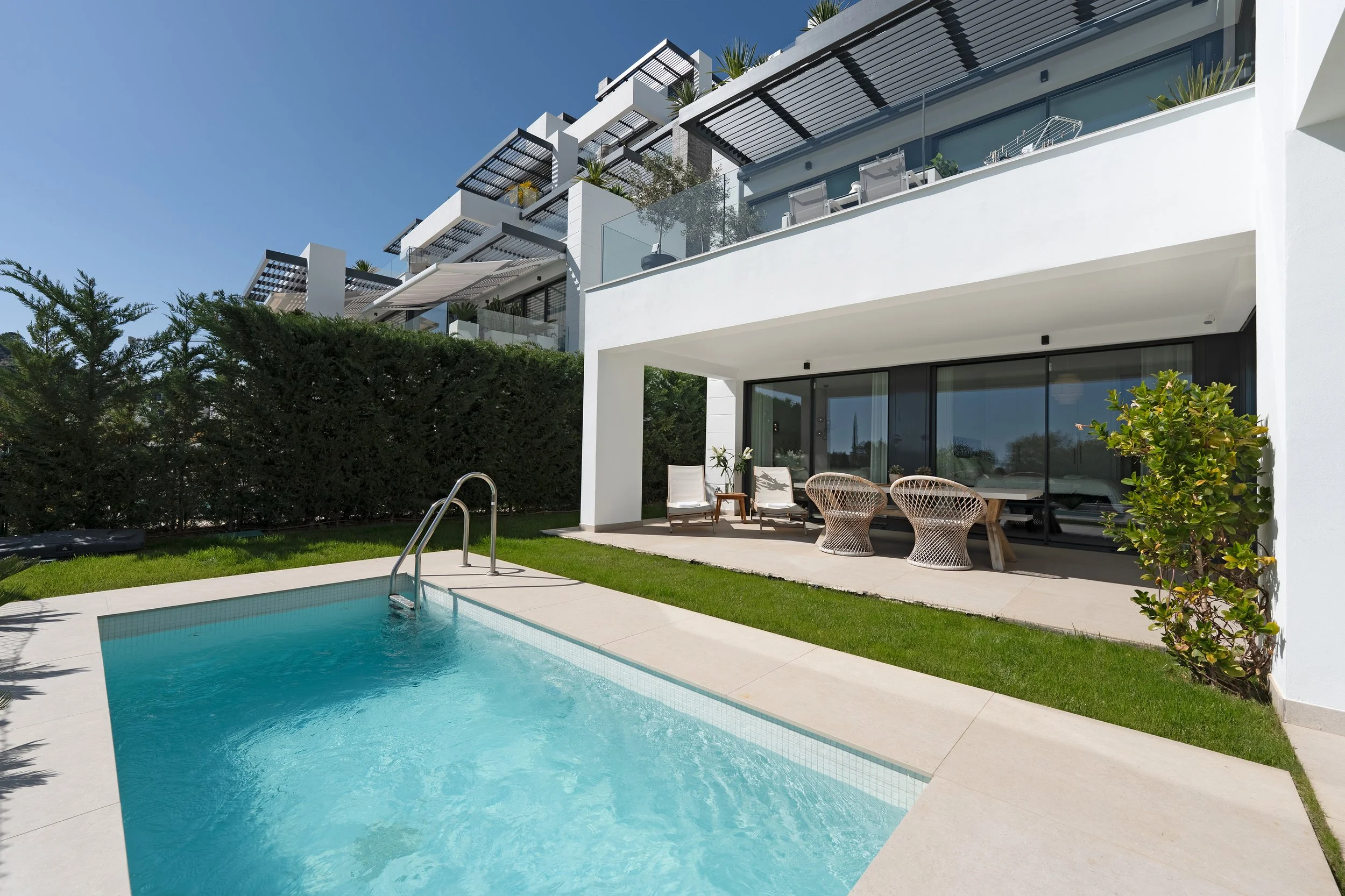 Modern apartment outdoor patio with swimming pool, seating area with chairs and table, and apartment building in the background.