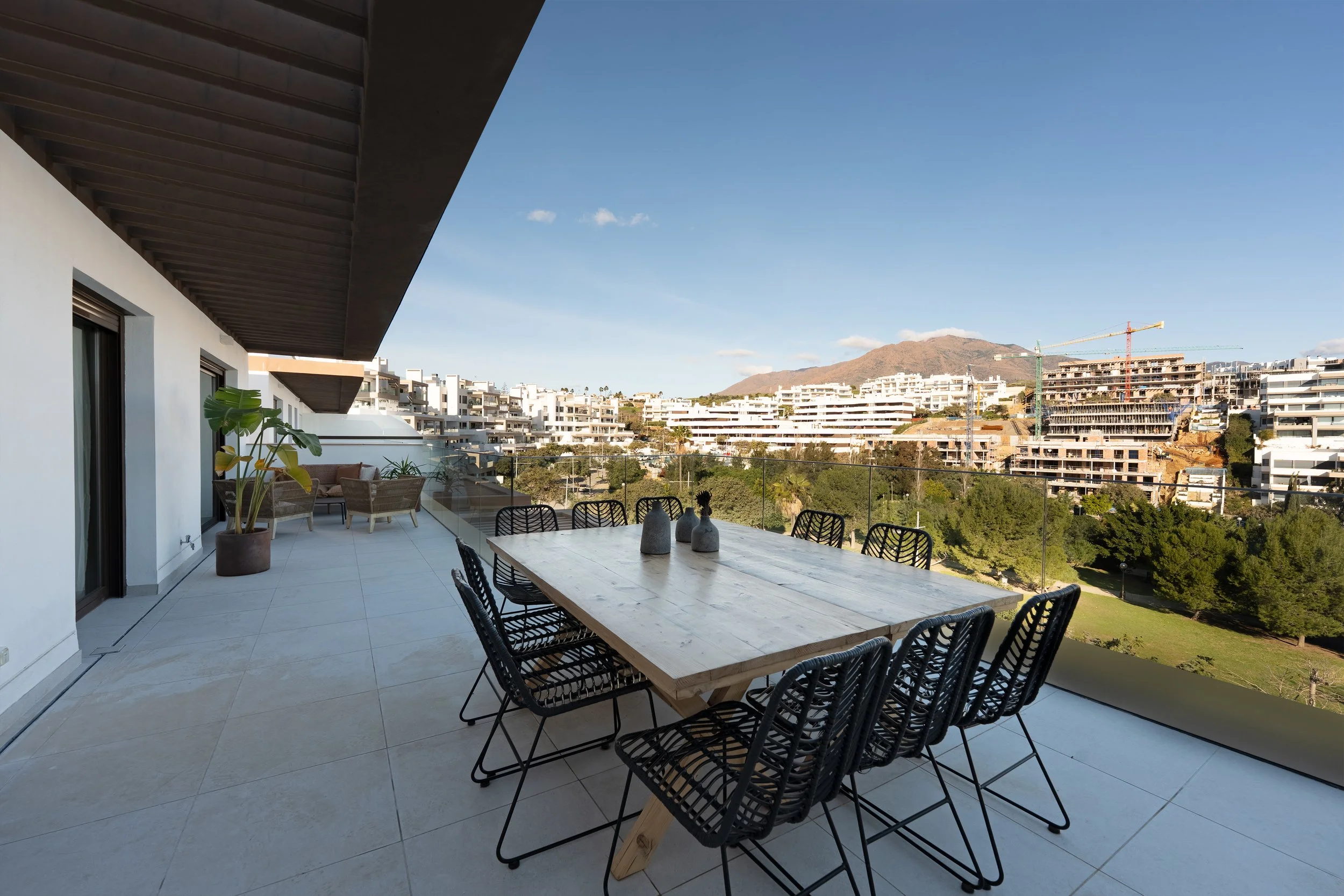Balcony with outdoor dining table, black chairs, potted plants, lounge seating, and cityscape view with construction cranes and distant mountains.