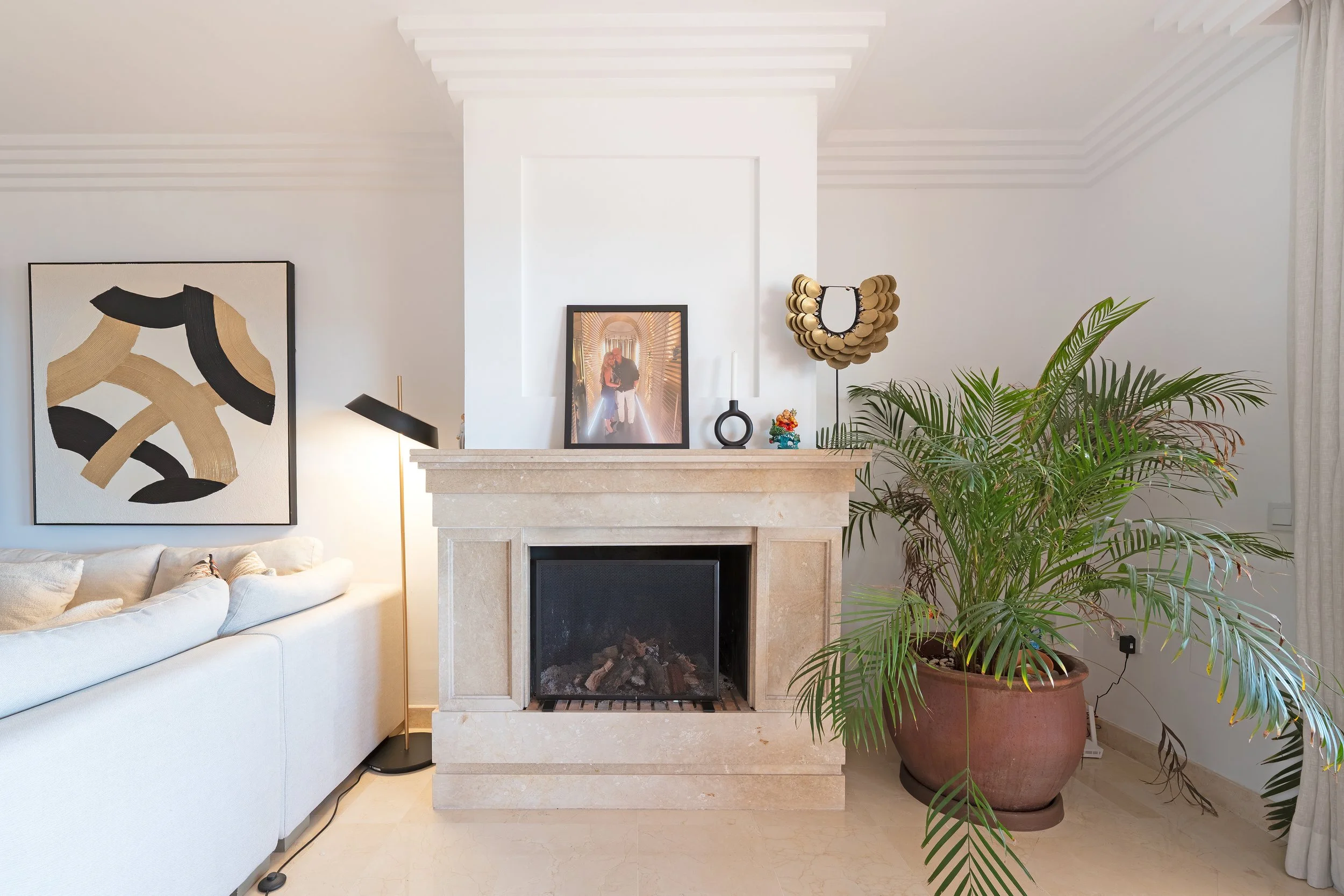 A living room with a white sofa, black and gold abstract art on the wall, a floor lamp, a fireplace with decorative items on the mantel, a large potted plant, and a white wall.