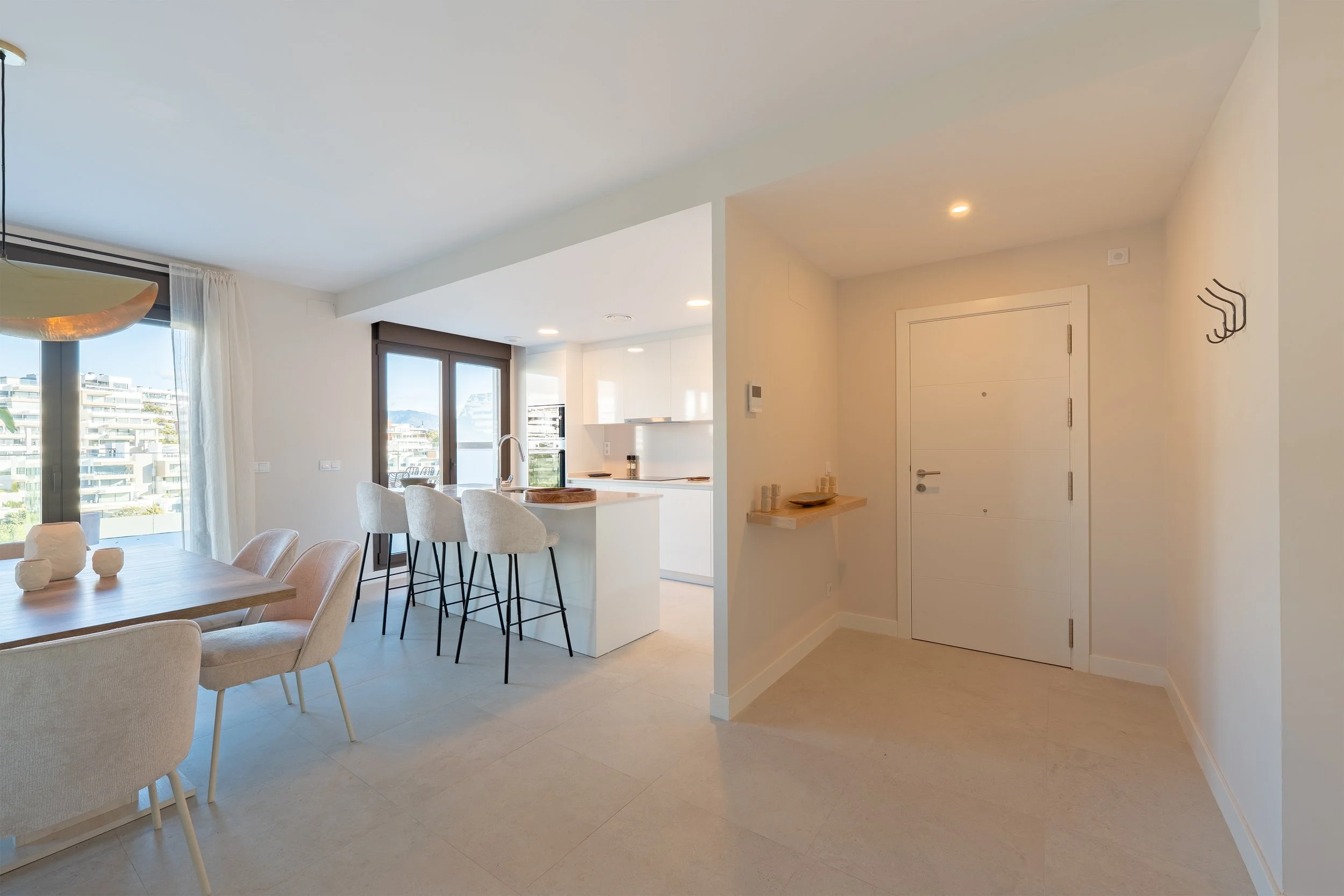 Open-concept living and dining area with a kitchen, barstools at a white island, dining table with chairs, and large windows with balcony view.