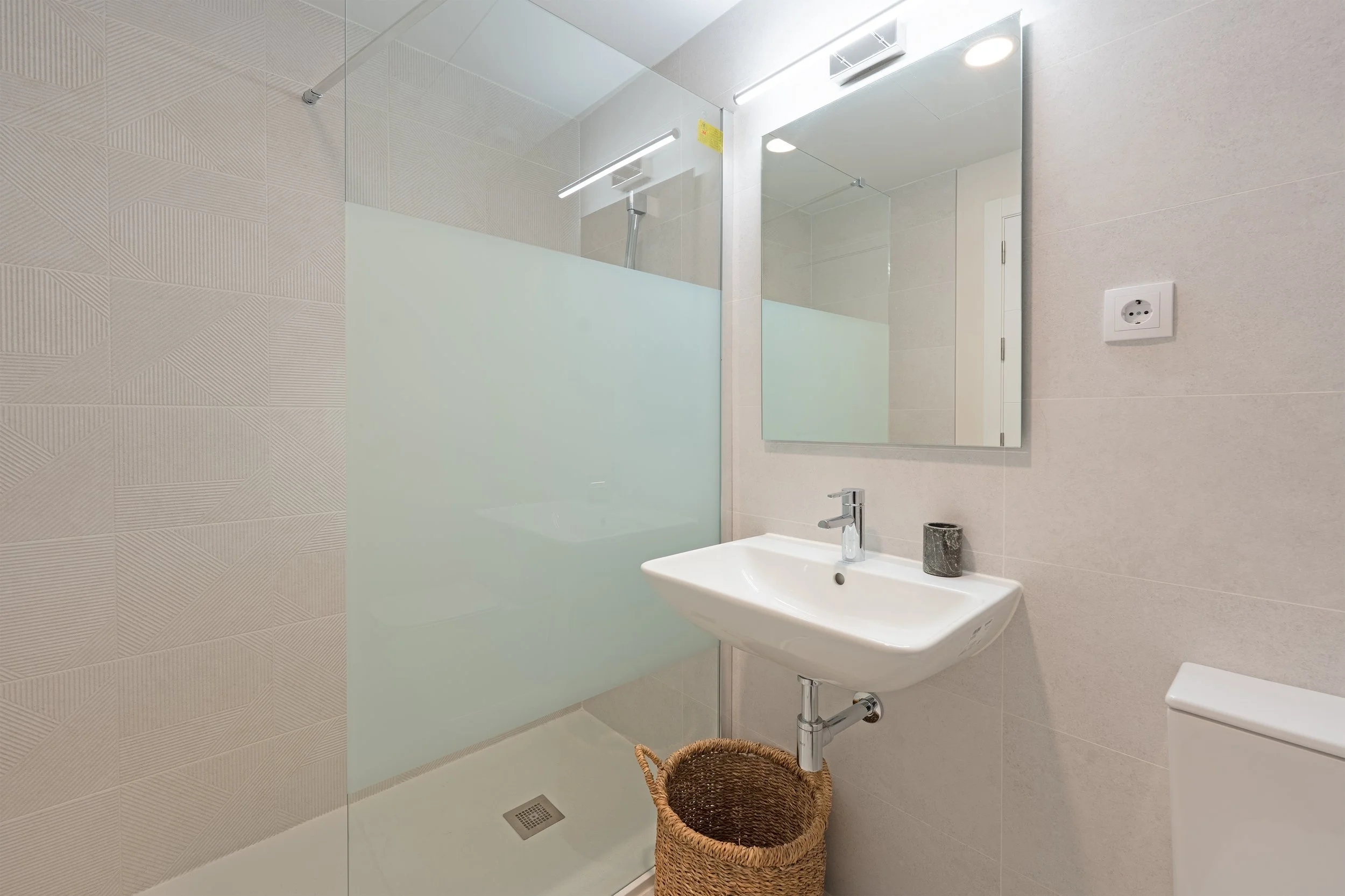 A modern bathroom with a shower area, a white sink with a faucet, a large mirror, a wicker basket underneath the sink, a soap dispenser, and a wall electrical outlet.