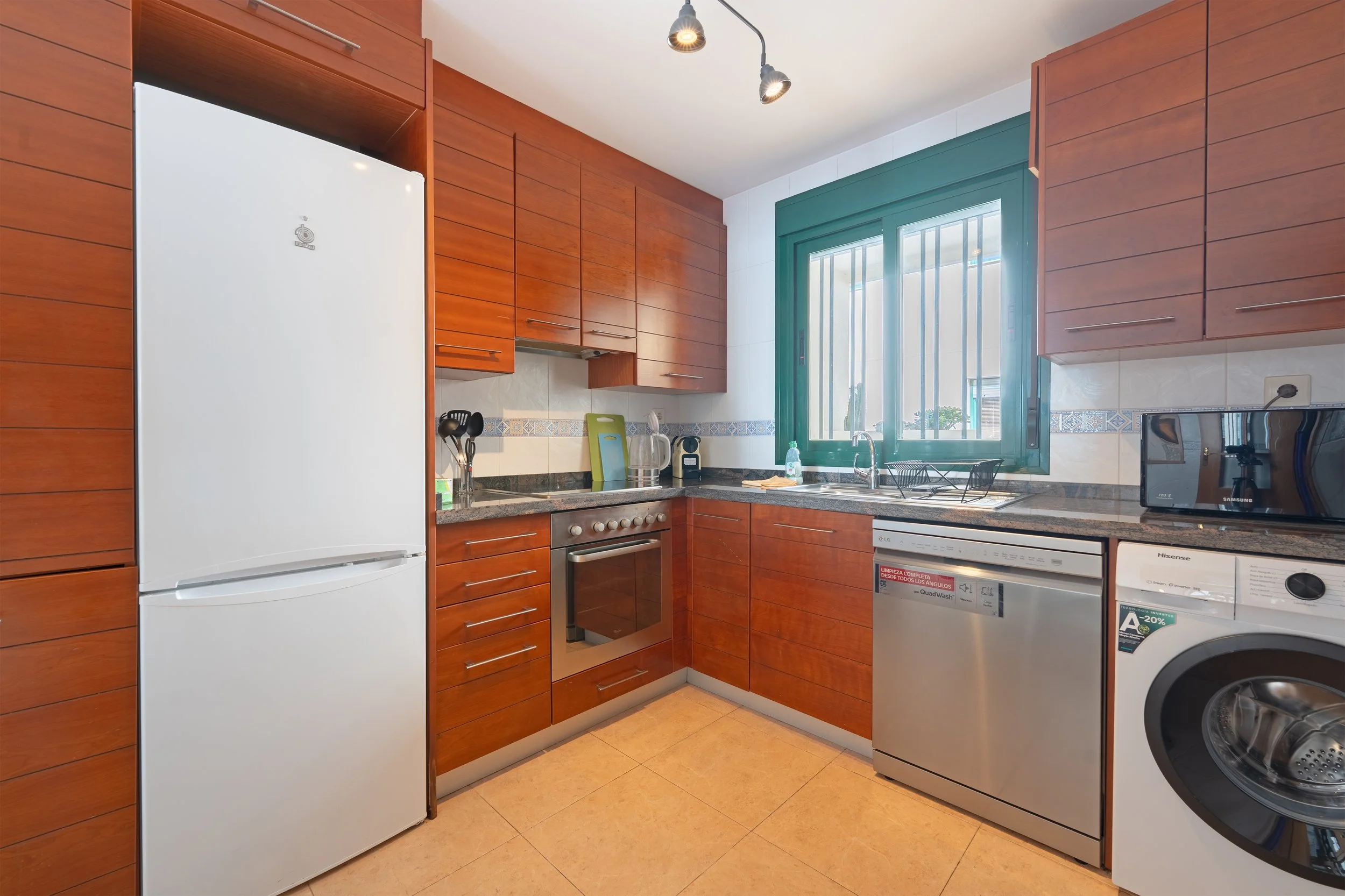 Kitchen with wooden cabinets, a white refrigerator, a stove, microwave, washing machine, and sink near a window with bars.