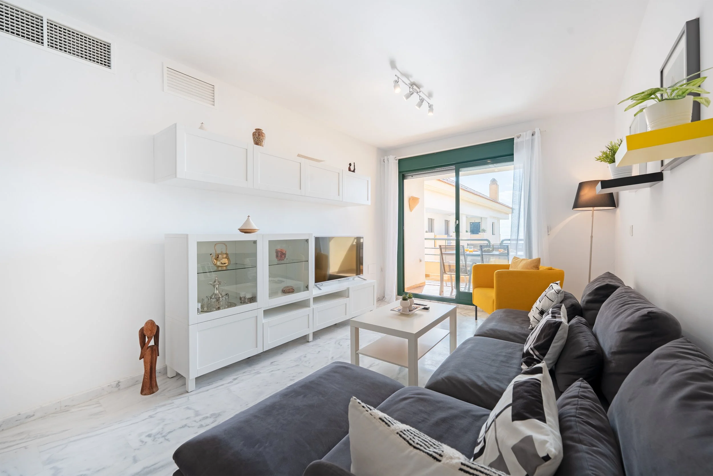 Bright living room with white walls and modern furniture, featuring a gray sofa with black-and-white pillows, a yellow armchair, a white coffee table, a white display cabinet, and sliding glass doors opening to a balcony with outdoor furniture.