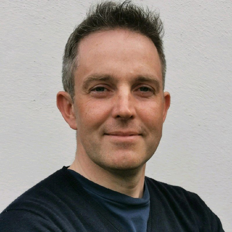 Portrait of a man with short dark hair, wearing a black shirt, standing in front of a plain white wall.