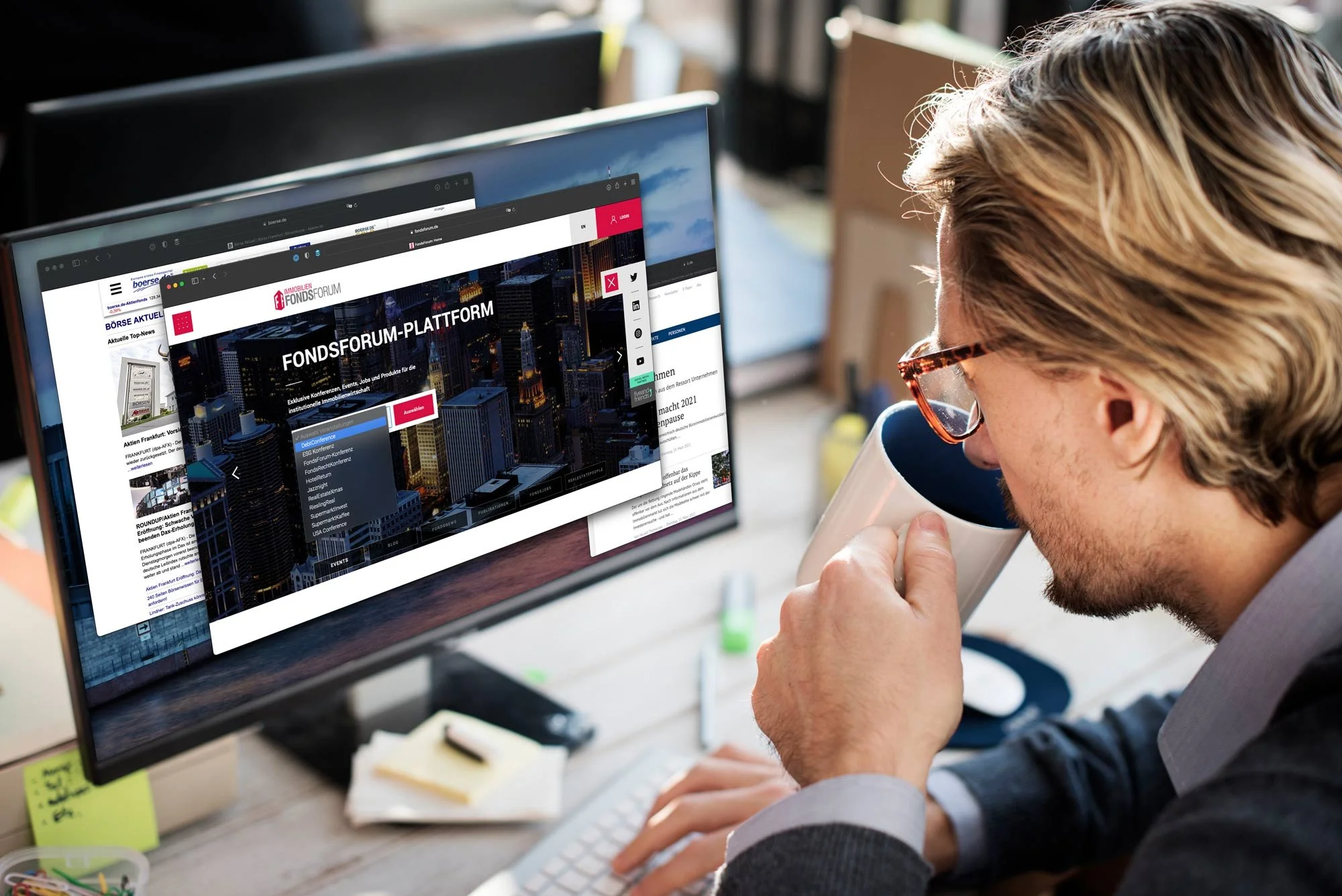 Ein Mann trinkt Kaffee während er an einem Computer arbeitet, der mehrere Webseiten zeigt, im Büro mit natürlicher Beleuchtung.