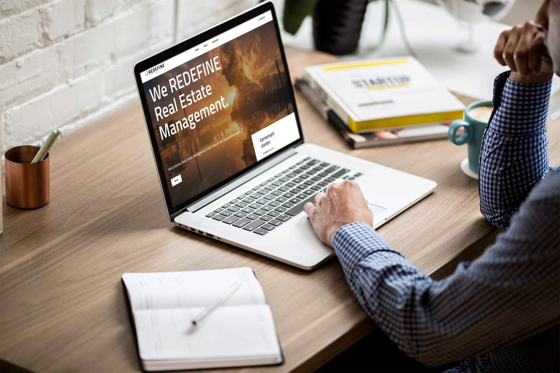Auf einem Schreibtisch arbeitet eine Person an einem Laptop, der eine Webseite mit dem Titel 'We REDEFINE Real Estate Management' zeigt. Im Hintergrund liegen Bücher und ein Kaffeetasse. Der Raum hat eine weiße Ziegelwand.
