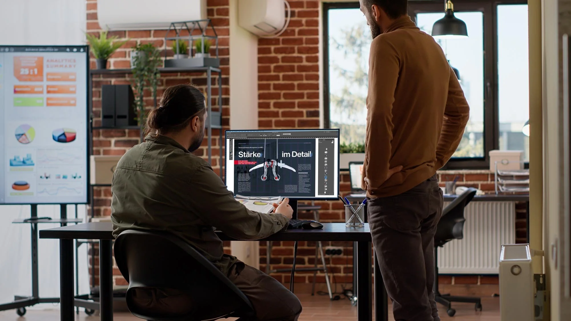 Zwei Männer arbeiten in einem modernen Büro mit braunem Holzfußboden und Backsteinwänden. Einer sitzt am Schreibtisch und sieht auf einen Bildschirm mit Grafiken und Text, der andere steht daneben. Im Hintergrund sind große Fenster, Pflanzen und weitere Büromöbel zu sehen.