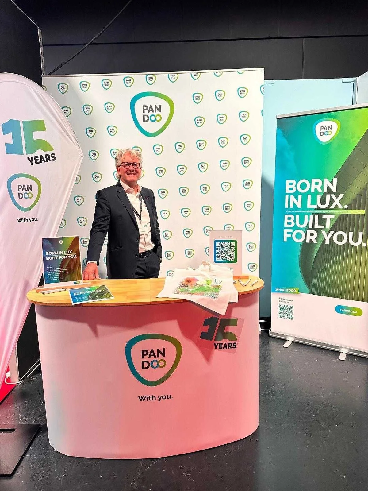 Ein Mann steht hinter einem Stand bei einer Messe oder Veranstaltung, mit Logos und Bannern von PanDoo, einem Unternehmen, das auf 15 Jahre Erfahrung zurückblicken kann. Der Stand ist mit bunten Bannern und Informationsmaterial decoriert.