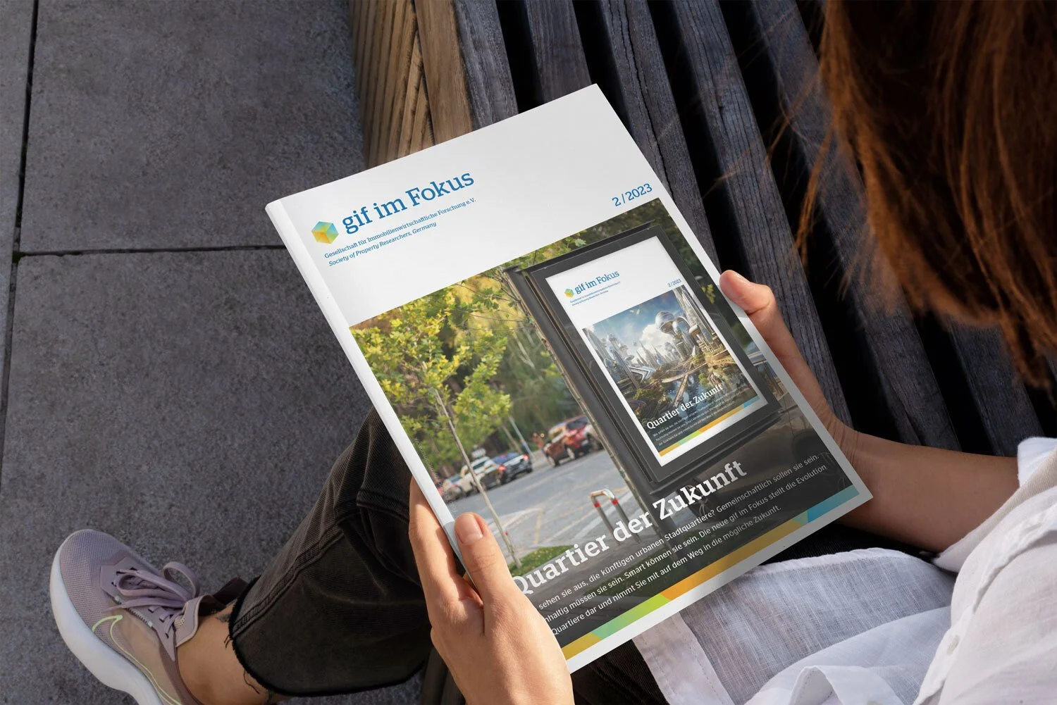 Zeitschrift mit dem Titel "gif im Fokus" liegt auf einem schwarzen Holztisch, Blick auf eine Straße mit Autos und grünen Bäumen, eine Person liest die Zeitschrift, die Person trägt weiße Schuhe, schwarze Hosen und ein weißes Oberteil. Auf dem Cover ist eine städtische Szene mit einem futuristischen Gebäude zu sehen.