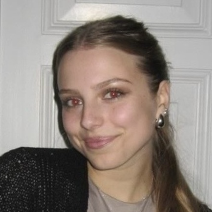 Ein junges Mädchen mit braunem Haar, das eine schwarze Jacke trägt und vor einer weißen Tür steht.