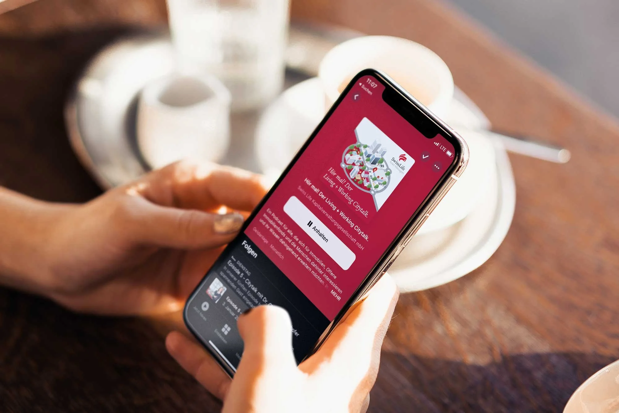 Eine Person hält ein Smartphone, das eine App mit einer rotem Hintergrund und einem Logo mit Gebäuden und Stadtillustration anzeigt, während sie in einem Café sitzt.